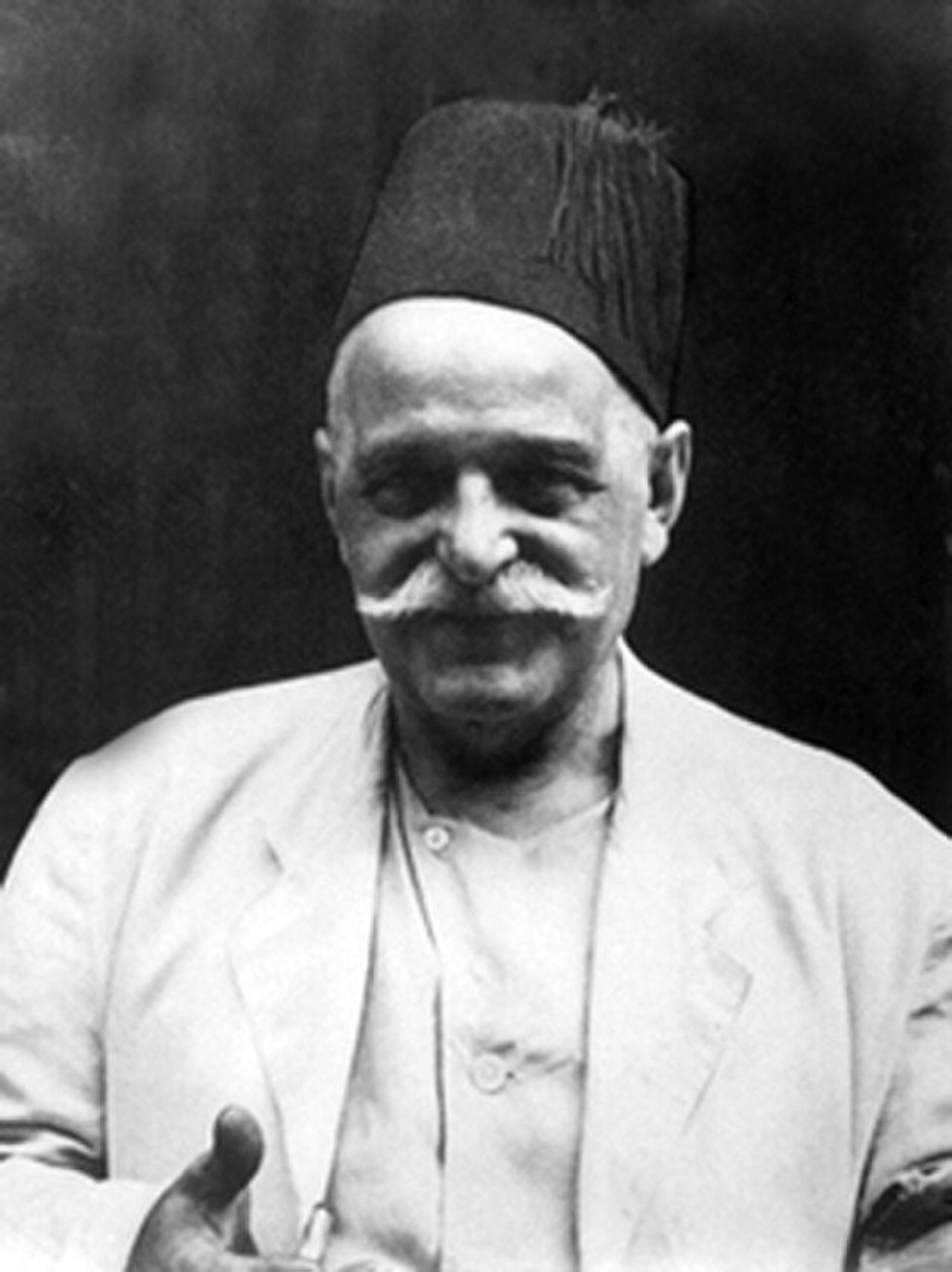 Black-and-white portrait of an older man with a white moustache and a dark fez, in a pale jacket, holding a hand toward the camera.