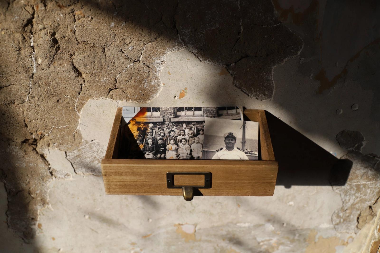 A small wooden card-catalogue drawer on a crumbling plaster wall, holding old black-and-white photographs.