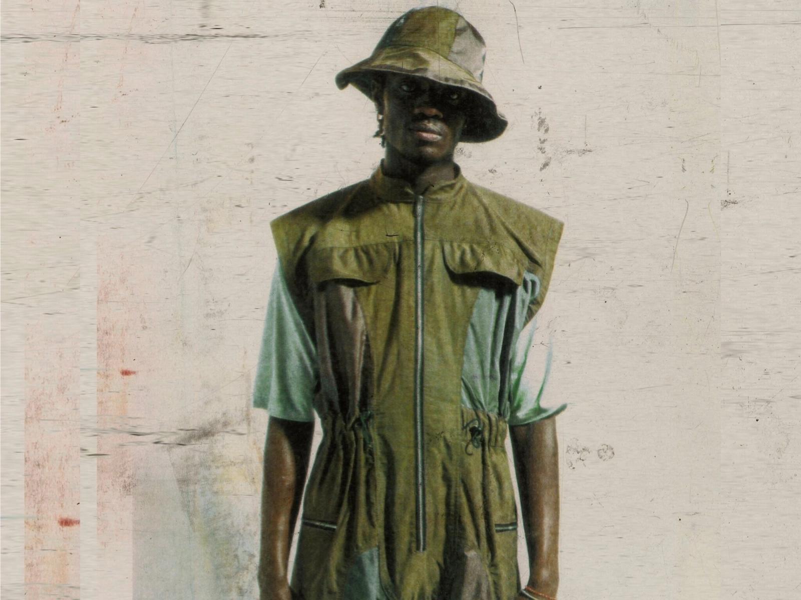 A model in an olive-green sleeveless jumpsuit and bucket hat stands against a scratched, pale backdrop.