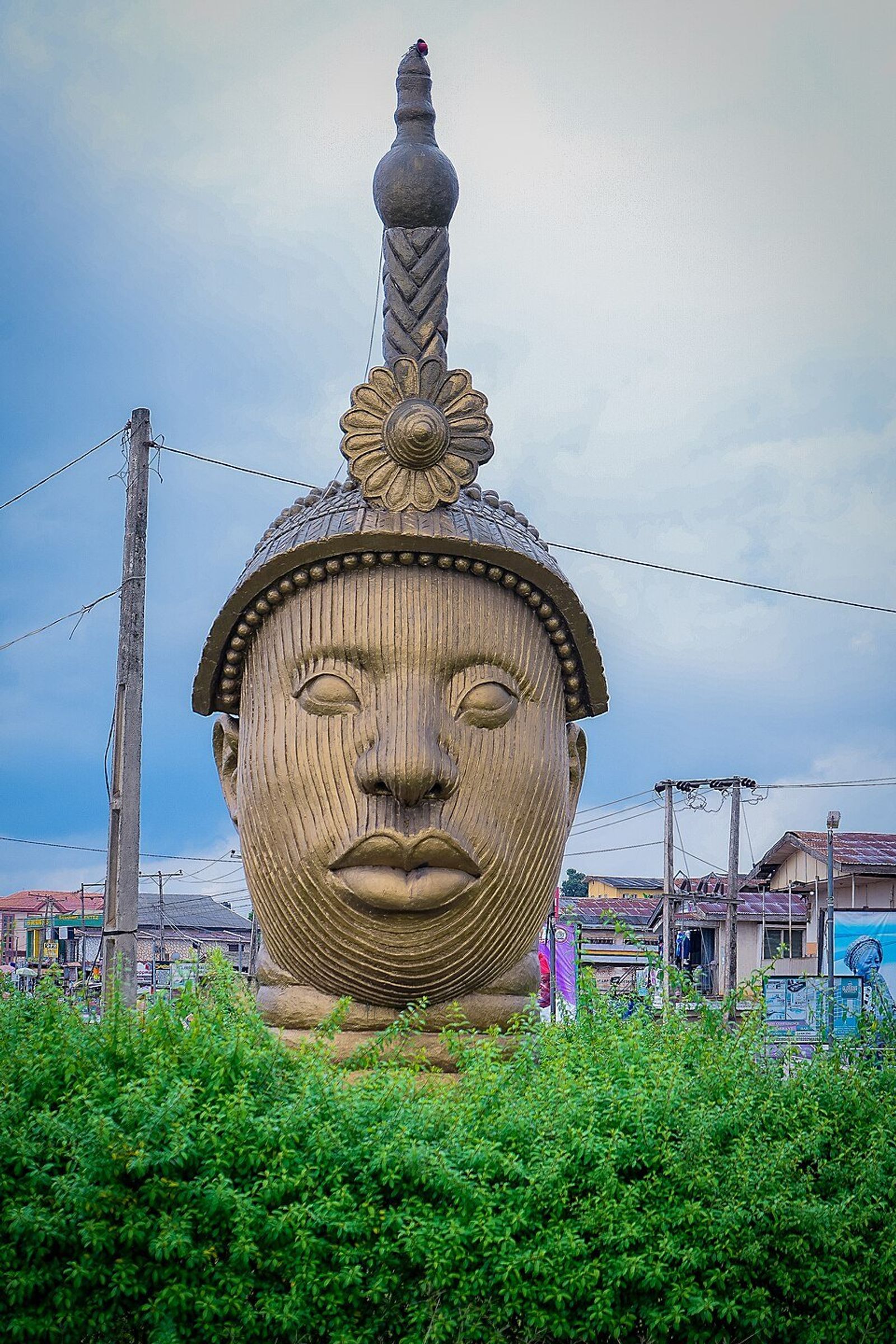 Monumental sculpture of a head wearing an ornate crown, rising above a green hedge against a blue sky and power lines.