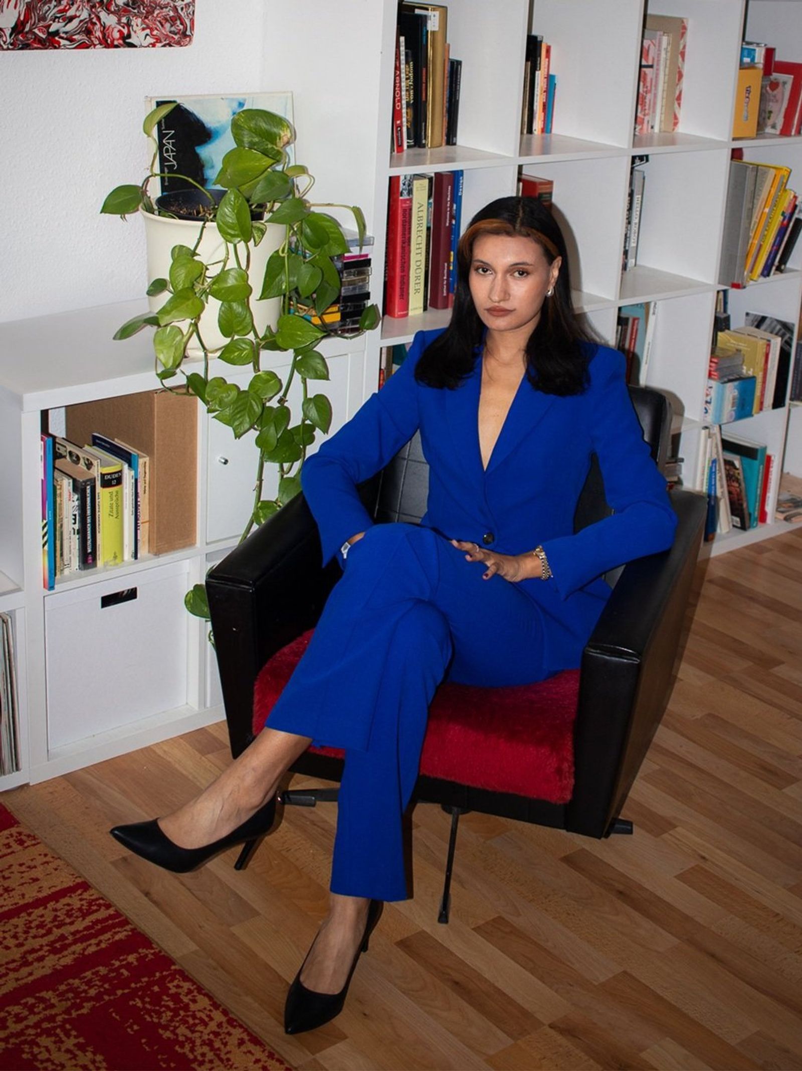 Jumoke Fernandez seated full-length in a black armchair, in a cobalt-blue suit, in a room with white bookshelves and a wooden floor.