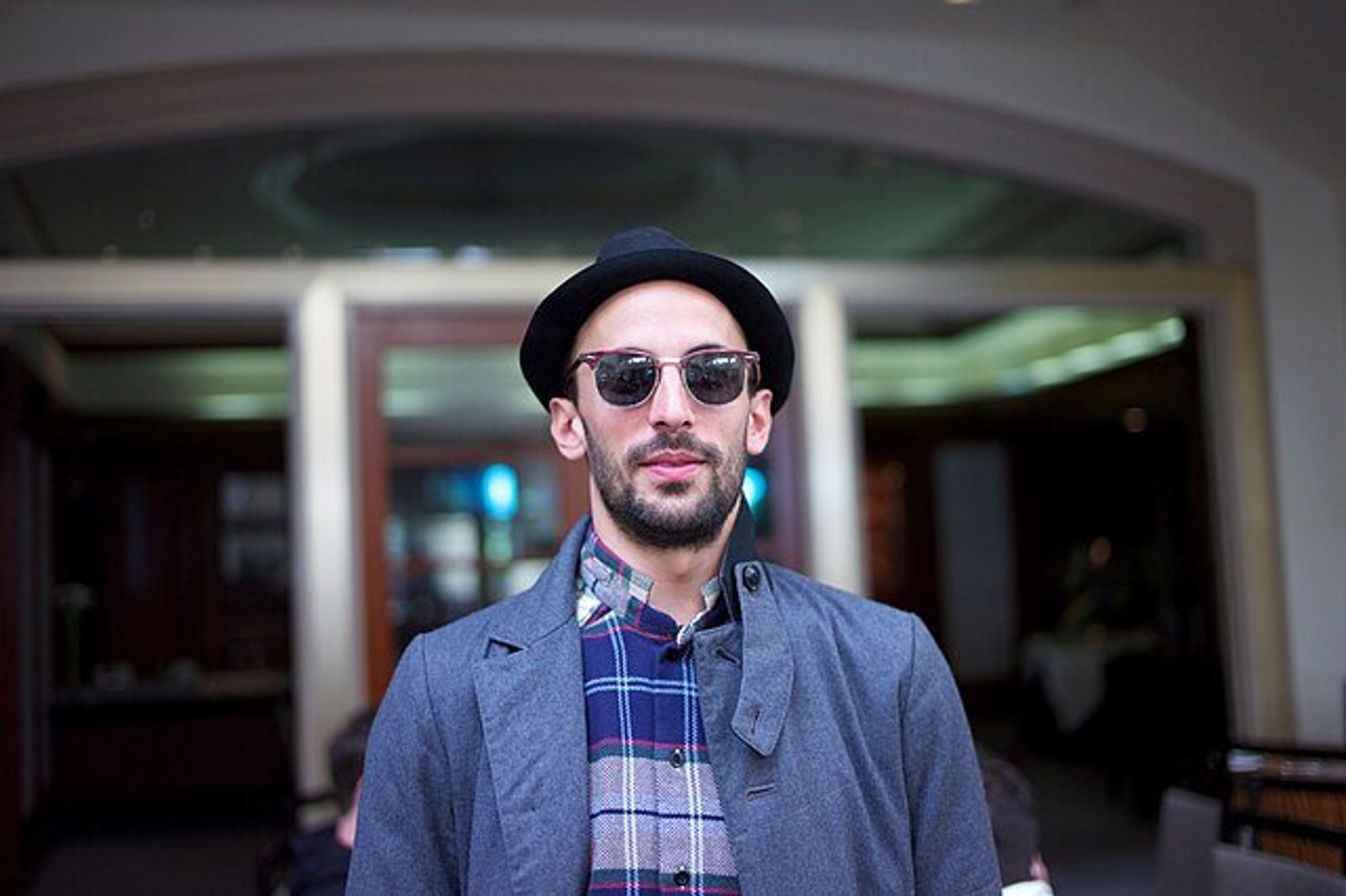 A bearded man in a black hat, dark sunglasses and a grey coat over a plaid shirt stands in an arched doorway.