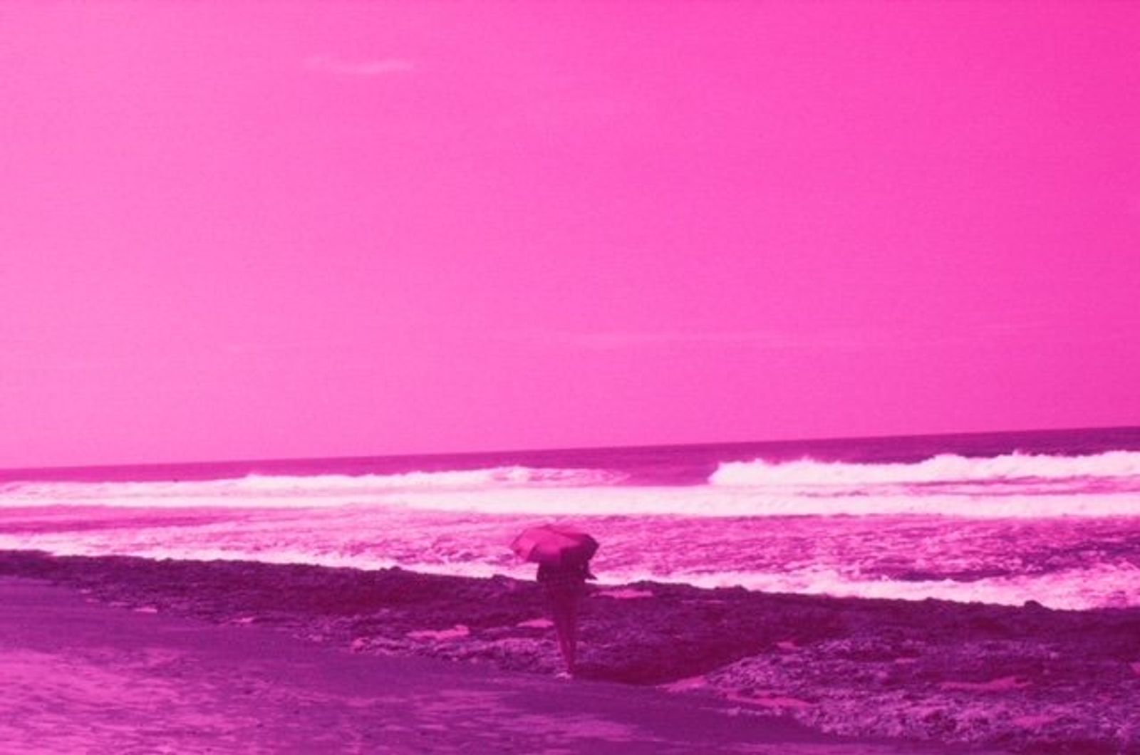 A figure bends over at the water's edge on a beach washed in magenta, with waves breaking beyond.
