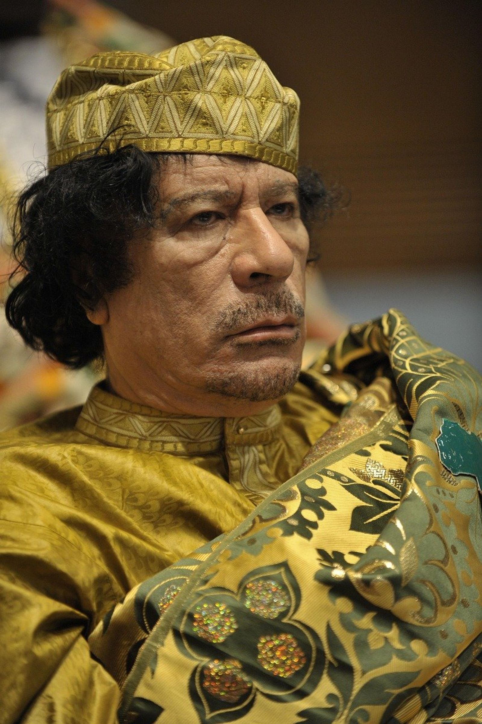 A man with dark curly hair in a gold embroidered cap and a gold patterned robe looks to one side with a stern expression.