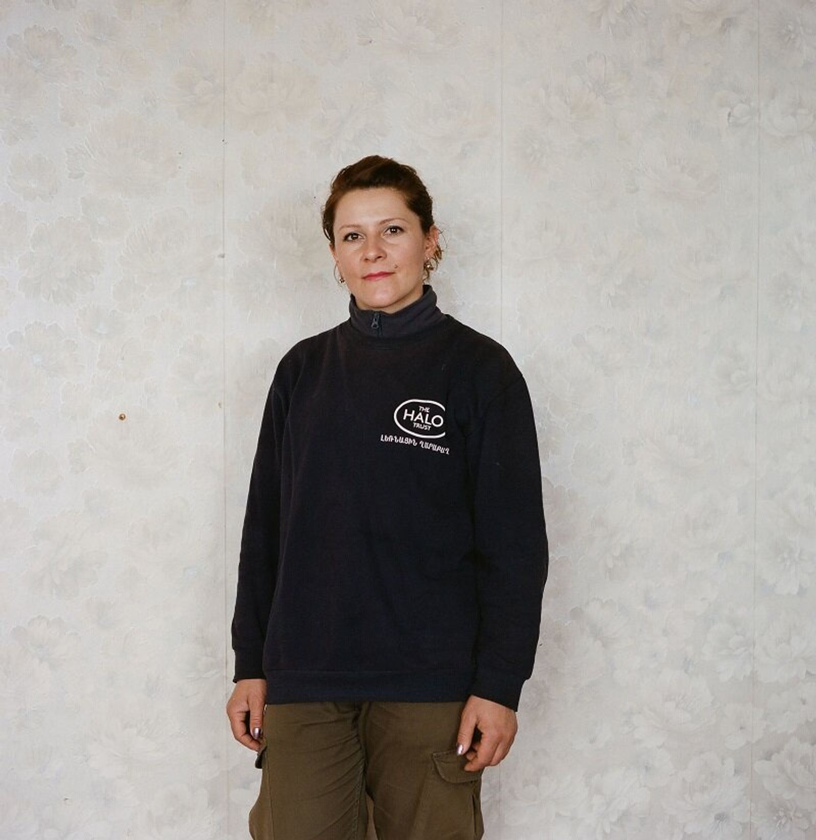 A woman with auburn hair pinned up, in a navy HALO Trust sweatshirt, stands against a pale wallpapered wall.