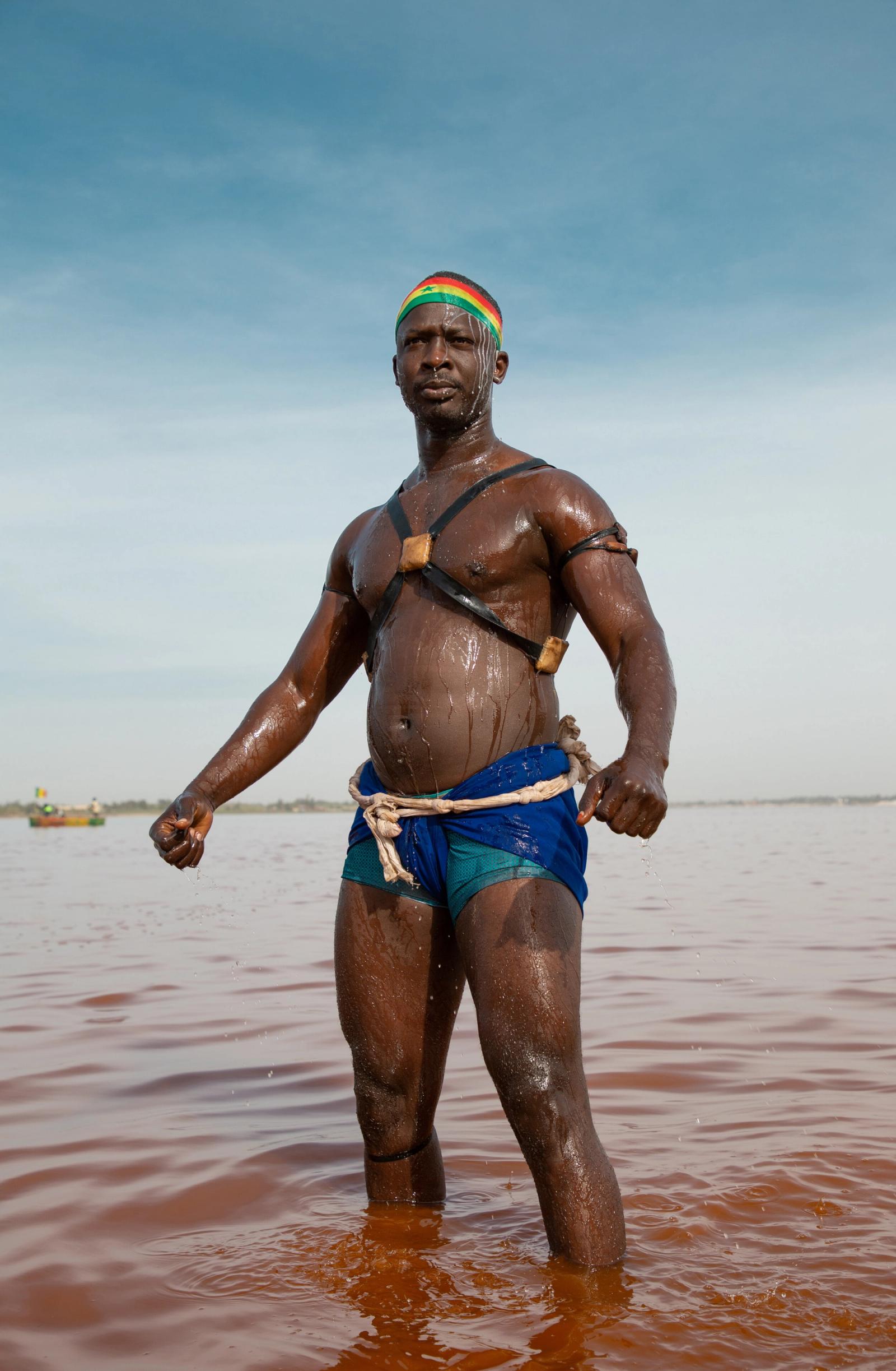A wrestler in a leather harness, rope belt and blue shorts stands waist-deep in a pink lake, a rainbow band round his head.
