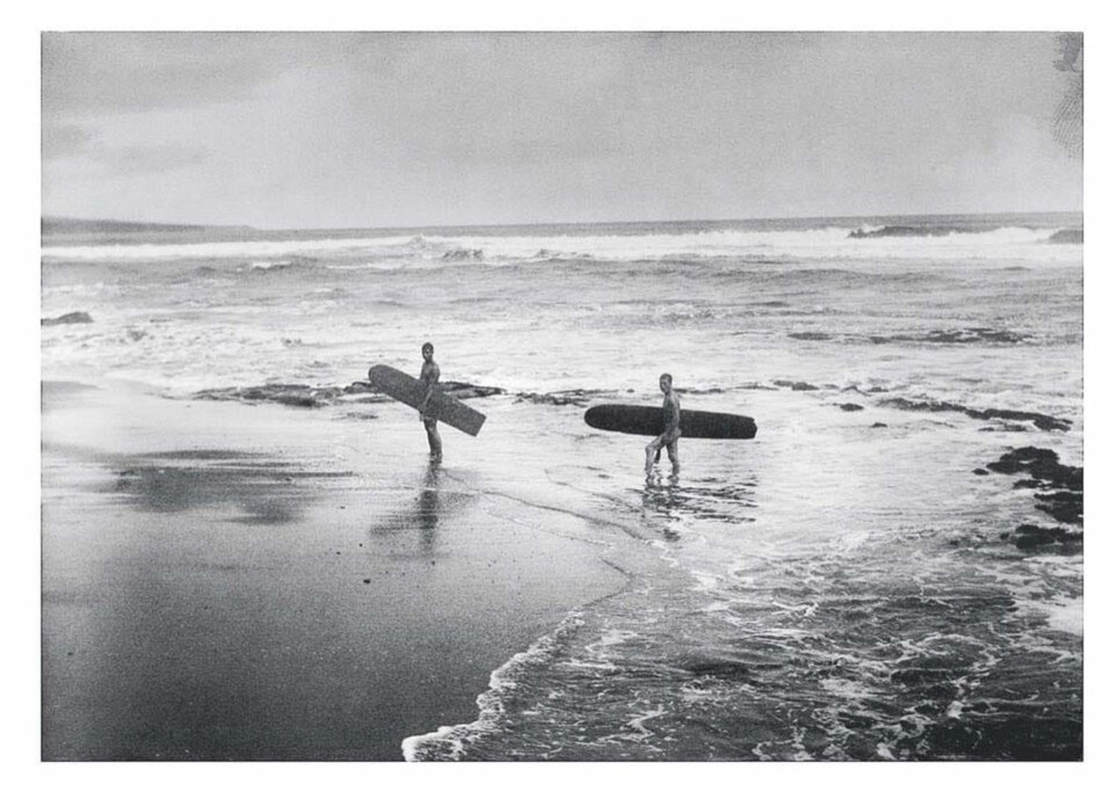 Black-and-white photograph of two surfers carrying longboards across wet sand as waves break behind them.