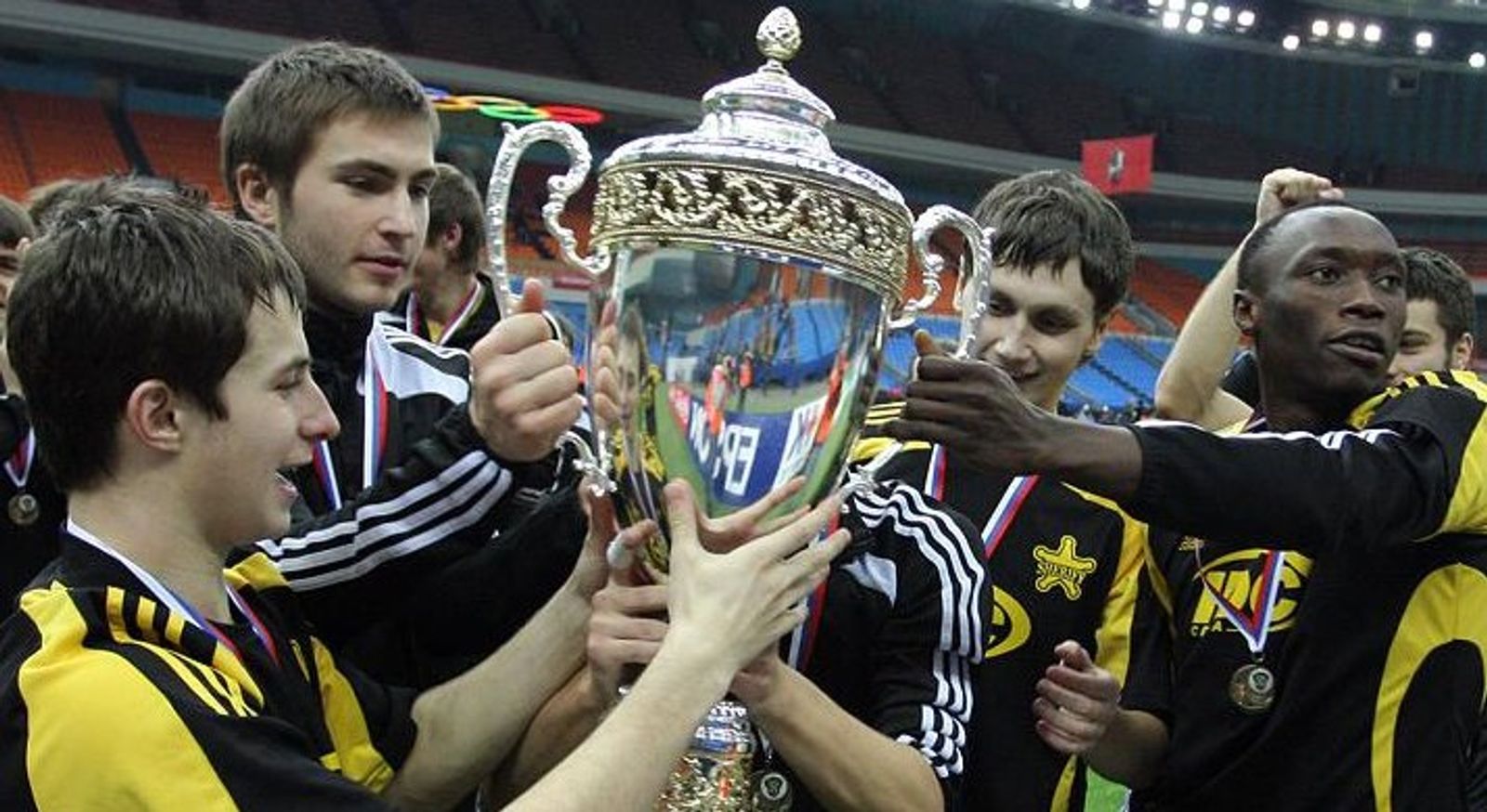 Players in yellow and black kits lift a large silver trophy together, medals around their necks.