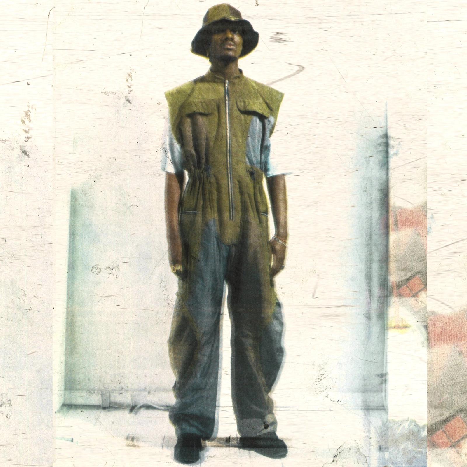 Full-length shot of a model in an olive jumpsuit with blue trousers and a bucket hat, against a scratched, pale backdrop.