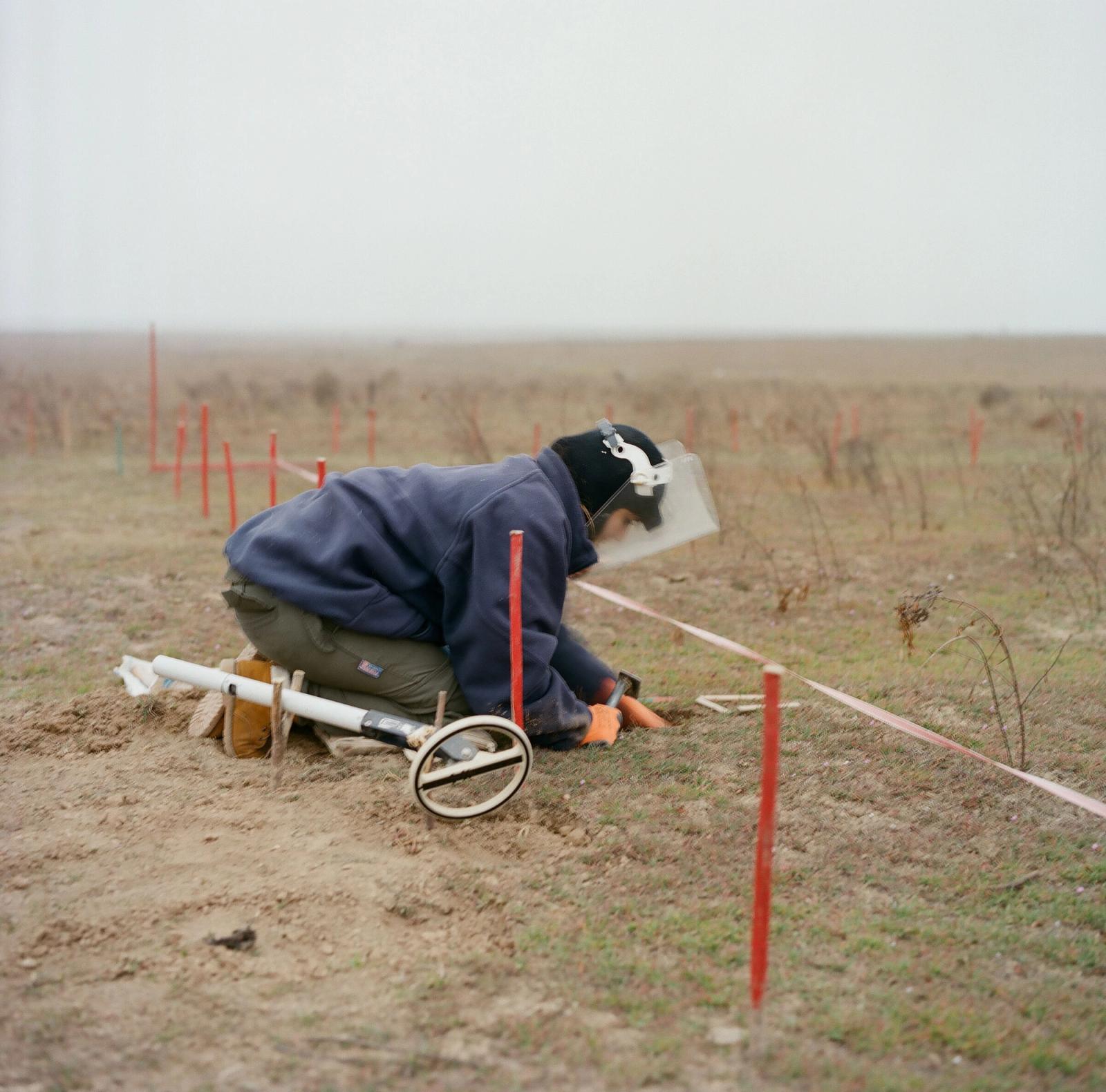A deminer in a visor and navy fleece kneels in an open field, probing the ground beside red marker stakes and tape.