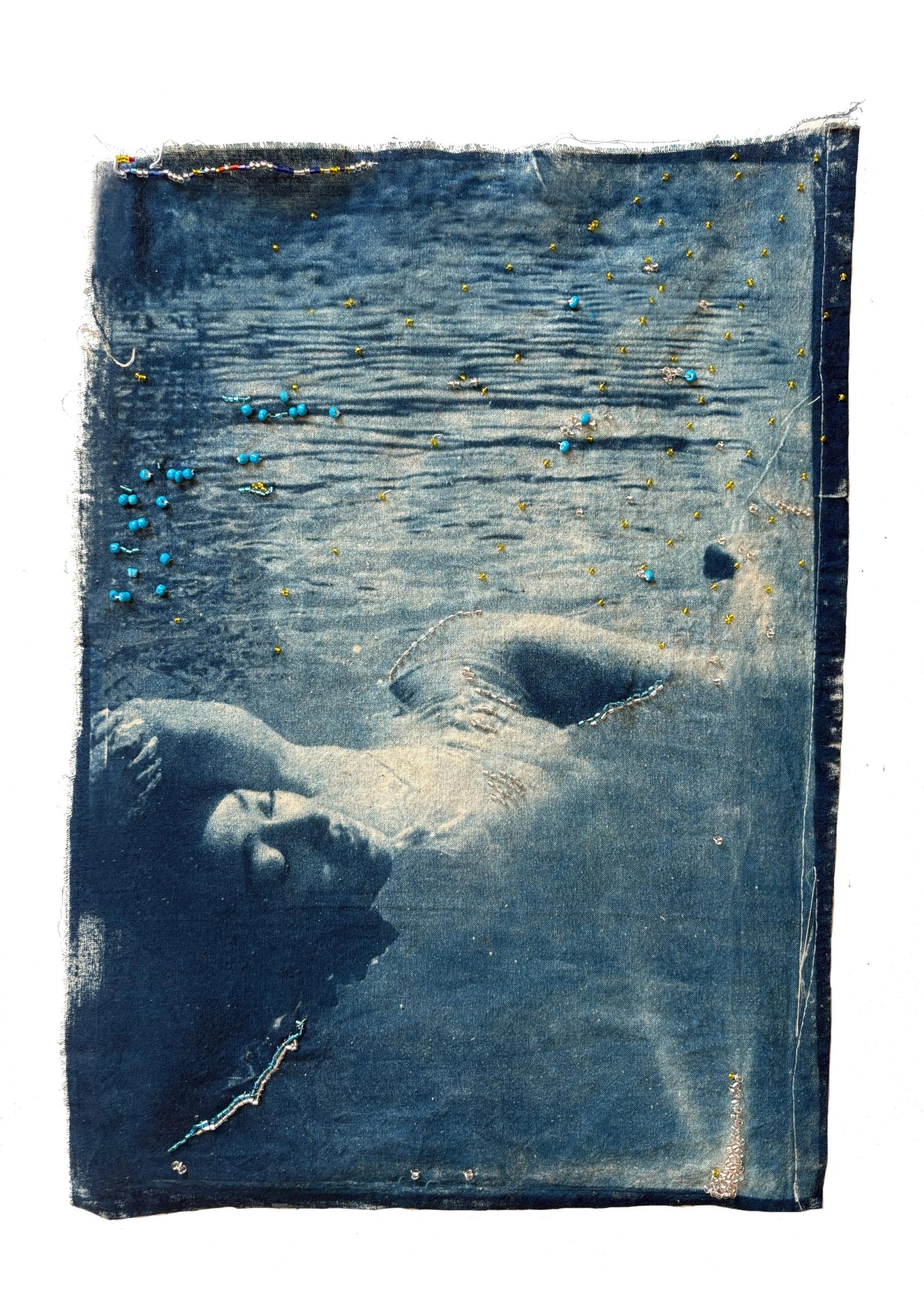 Cyanotype print on stitched fabric showing a woman's face and shoulders half-submerged in blue water, scattered with small beads.