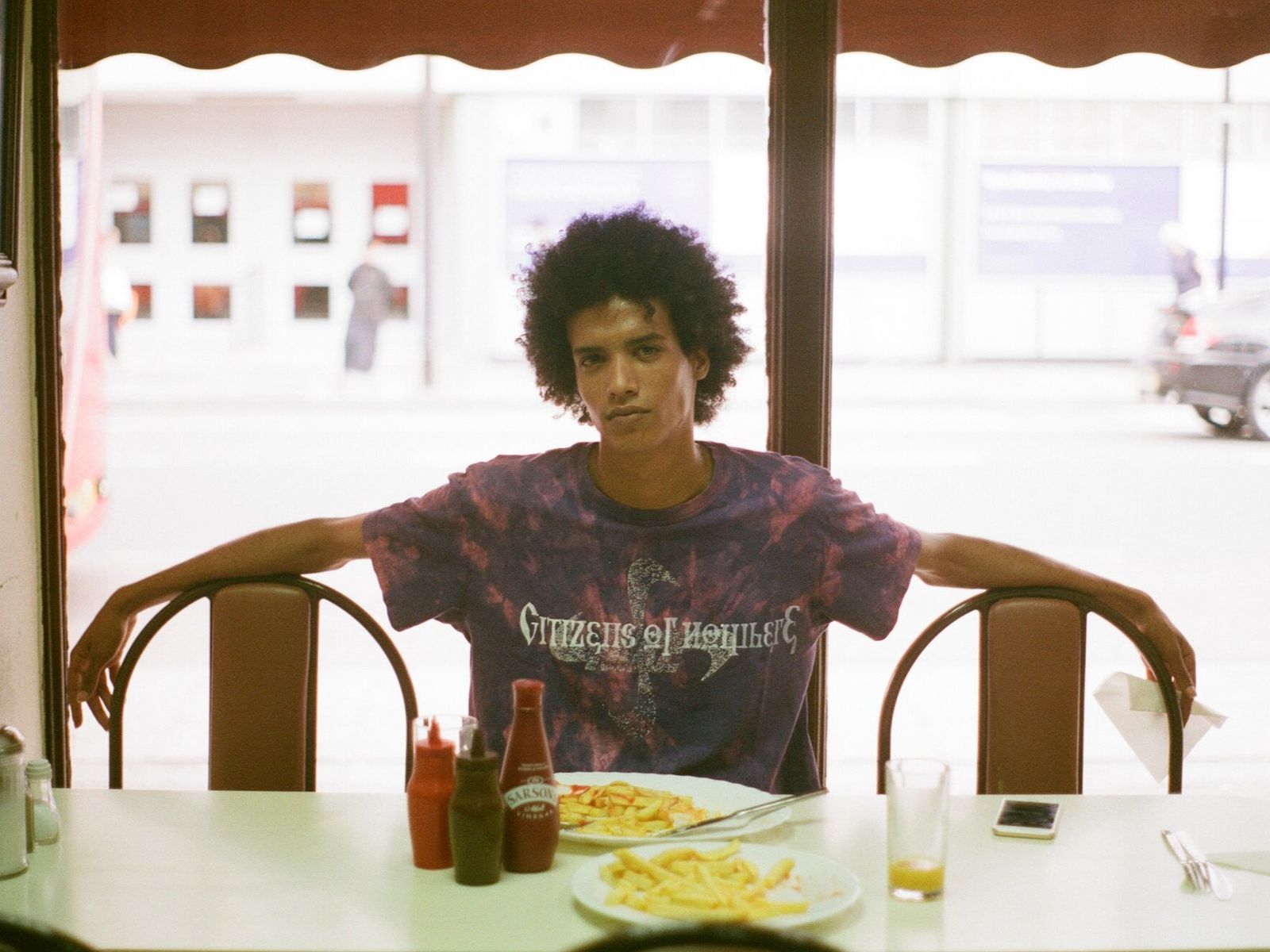 A young man with curly hair in a purple tie-dye Citizens of Nowhere t-shirt sits at a diner table with chips, arms on the chair backs.