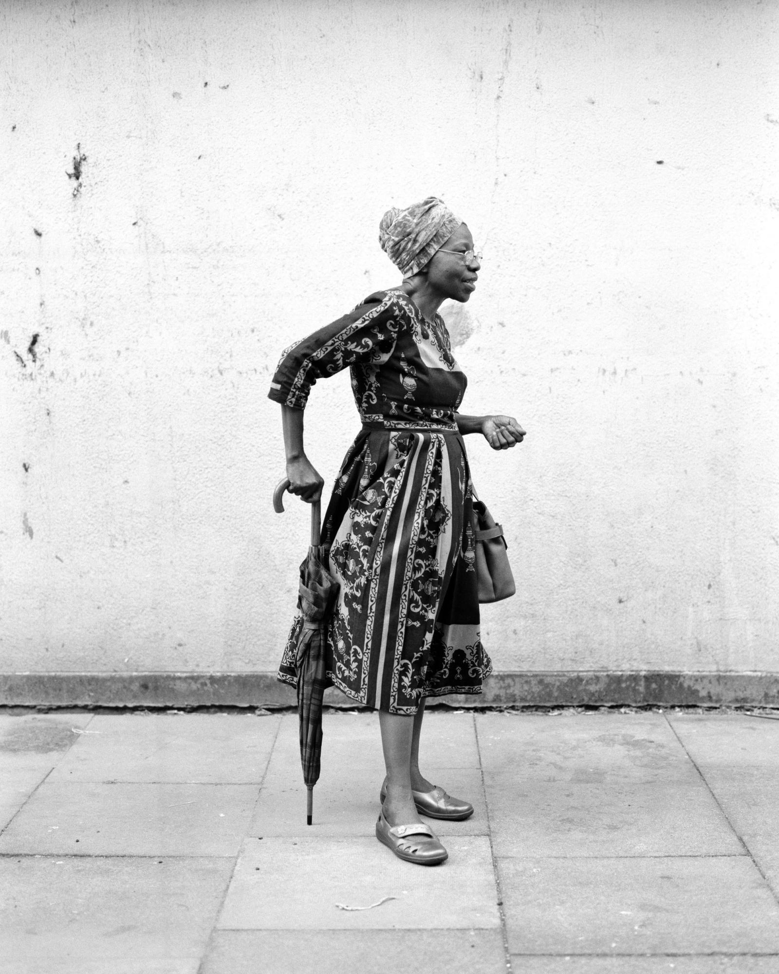 Black-and-white portrait of an older woman in a headwrap, glasses and patterned dress, holding a folded umbrella against a pale wall.
