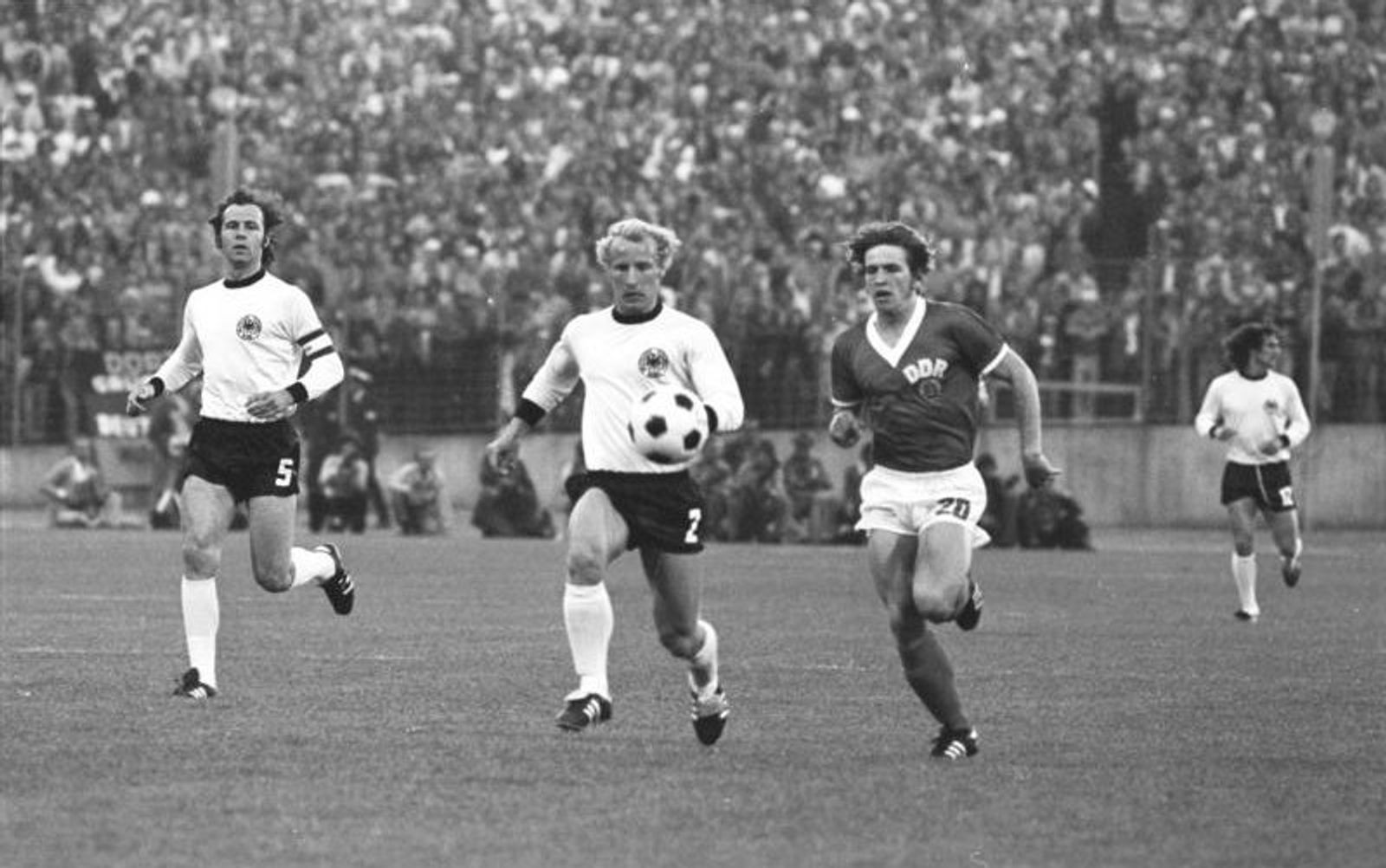 Black-and-white photograph of a match between West and East Germany: a player in white controls the ball as a DDR-shirted player chases him.