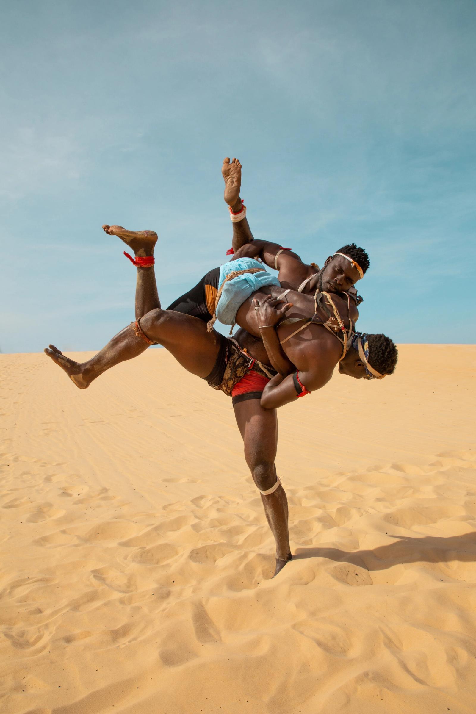 Two wrestlers on a sand dune, one lifting the other upside down mid-throw against a blue sky.