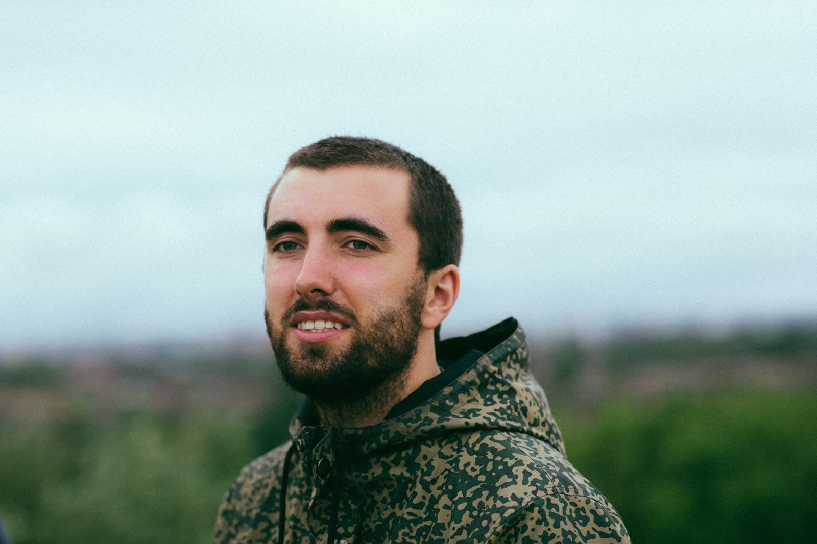 Milo Blake, smiling, with a short beard and a camouflage-print hoodie, against a cloudy sky and blurred green hills.