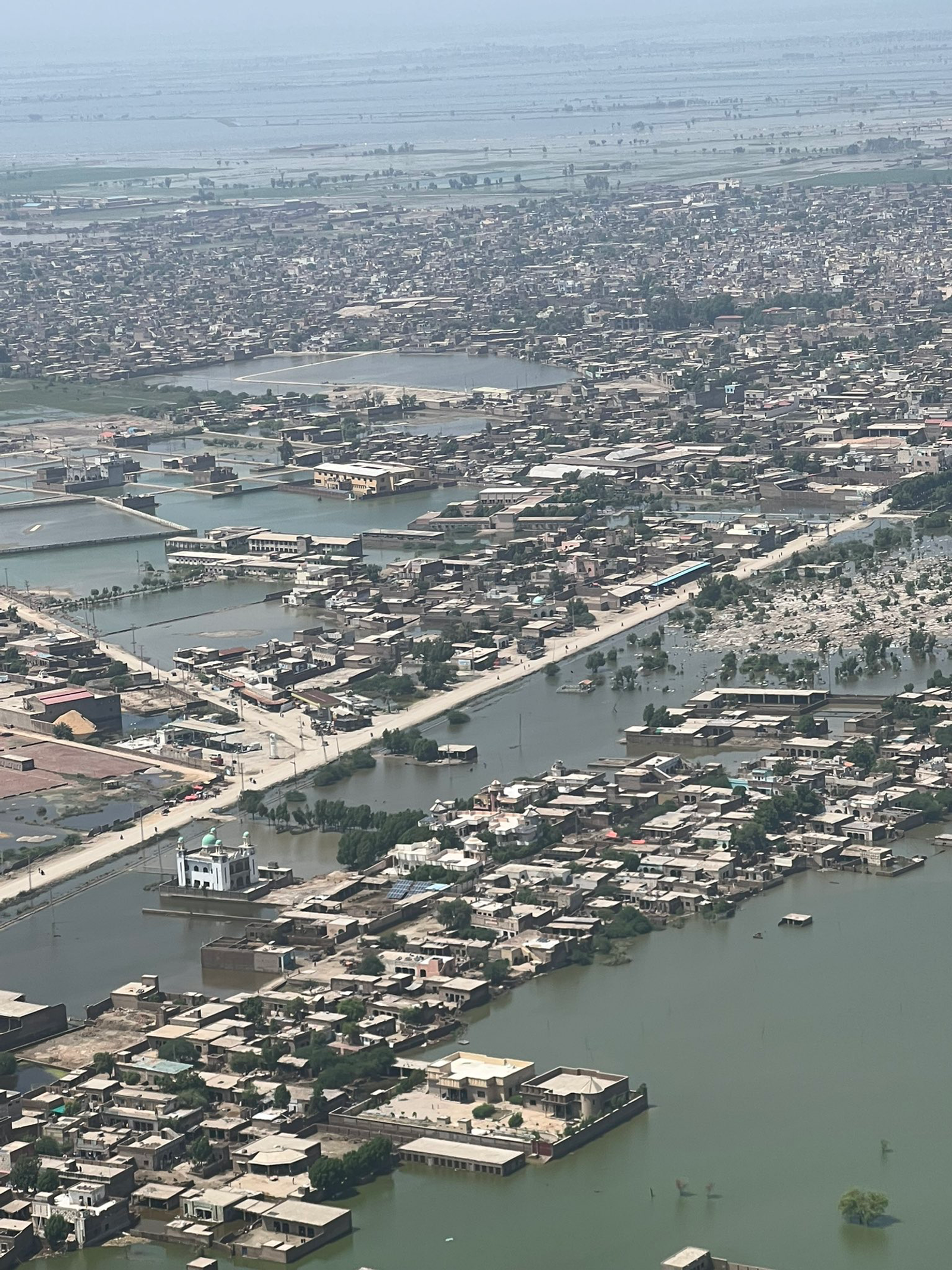 Aerial view of a flooded town in Pakistan, with roads and rooftops standing among brown-green floodwater.