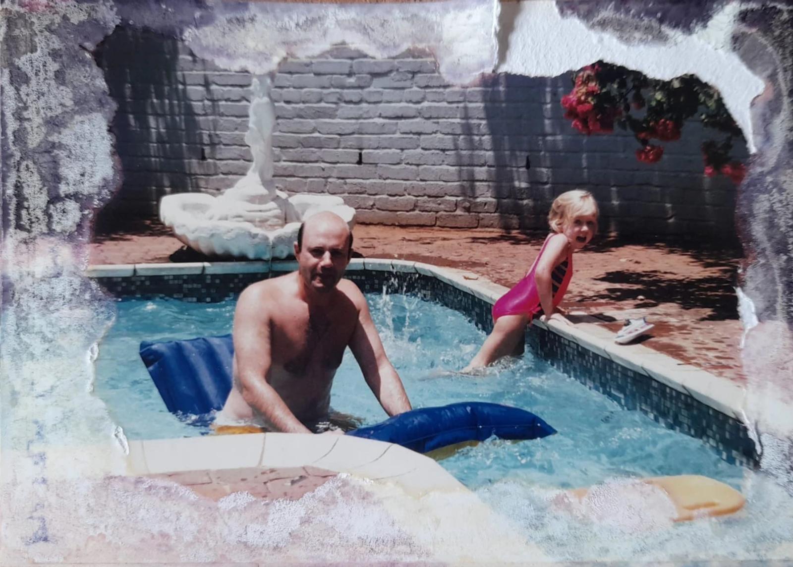 Water-damaged family photograph: a man stands in a backyard swimming pool holding a blue lilo while a small girl in a pink swimsuit sits on the edge. Damp has eaten the print's edges into white and violet blooms.