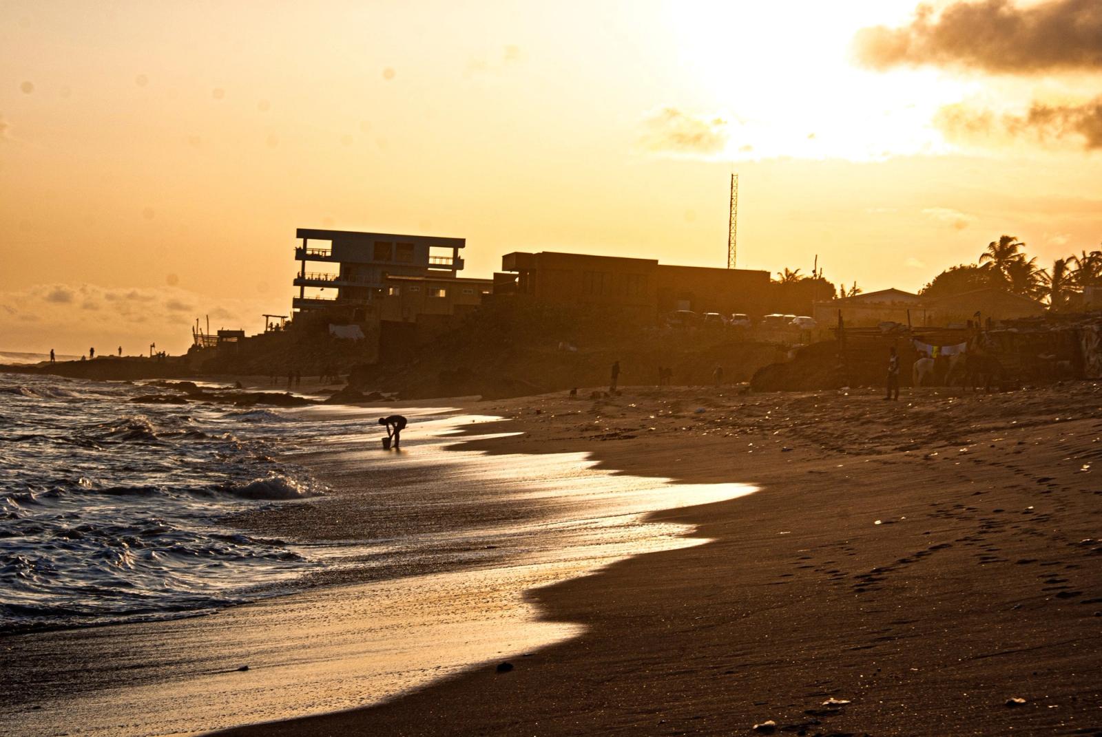 Sunset over a dark-sand beach, with silhouetted buildings along the shore and a lone figure crouching at the water's edge.