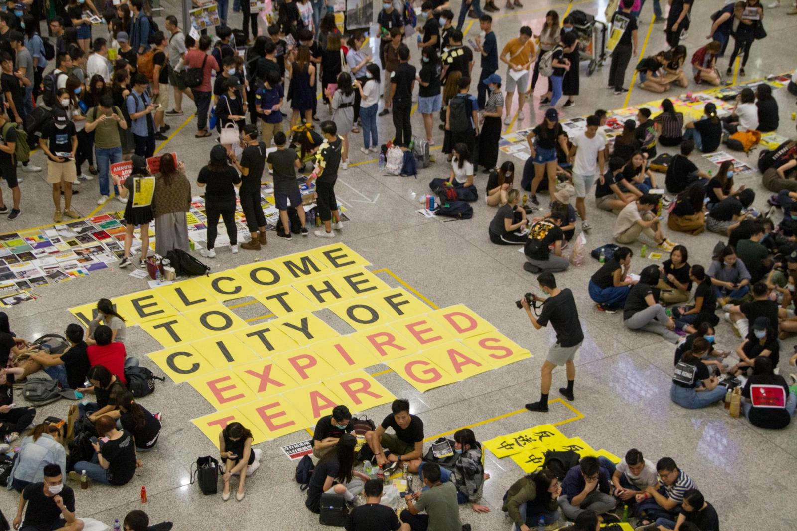 Protesters in black sit on an airport concourse floor around a yellow banner reading "Welcome to the city of expired tear gas".