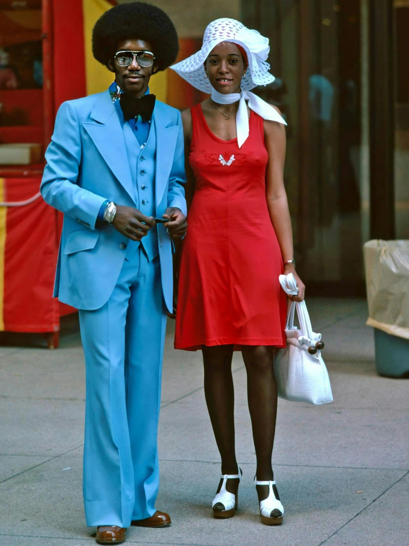 A man in a pale-blue suit, bow tie and glasses stands arm in arm with a woman in a red dress and white sun hat on a pavement.