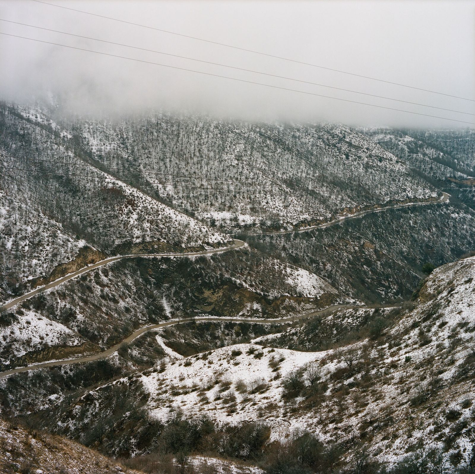 A winding road climbs through snow-dusted mountains under low cloud, with power lines across the sky.