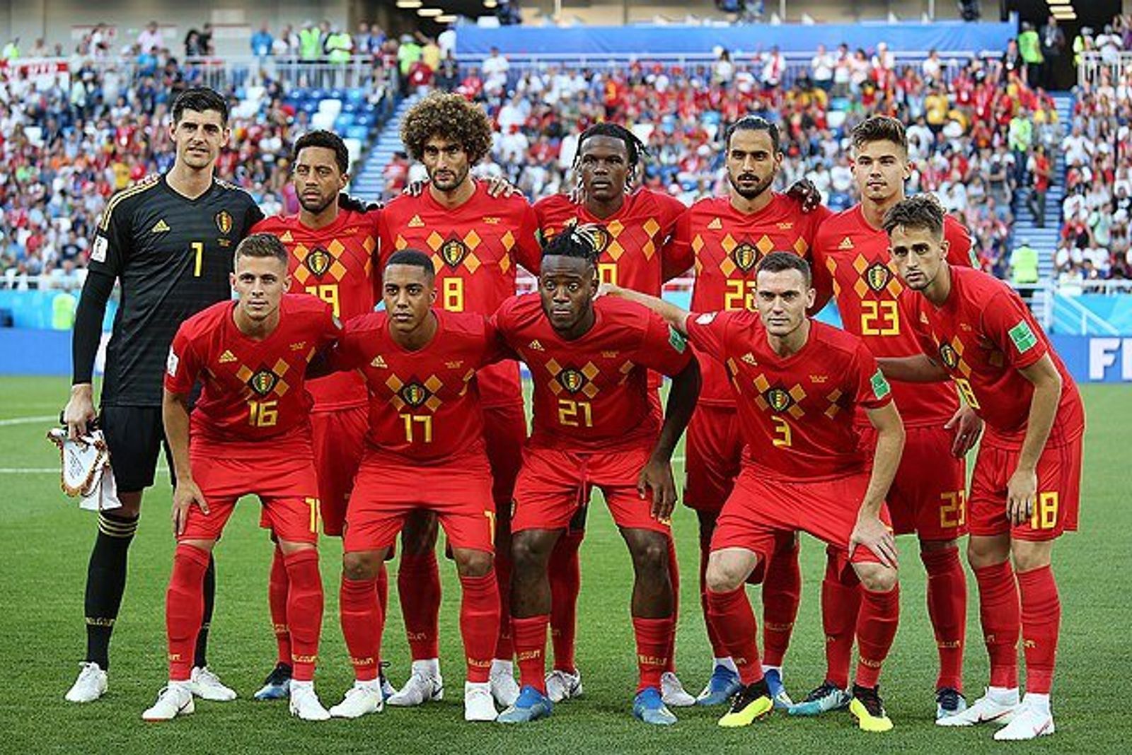 Belgium's players in red kits pose in two rows on the pitch before a match, with a stadium crowd behind.