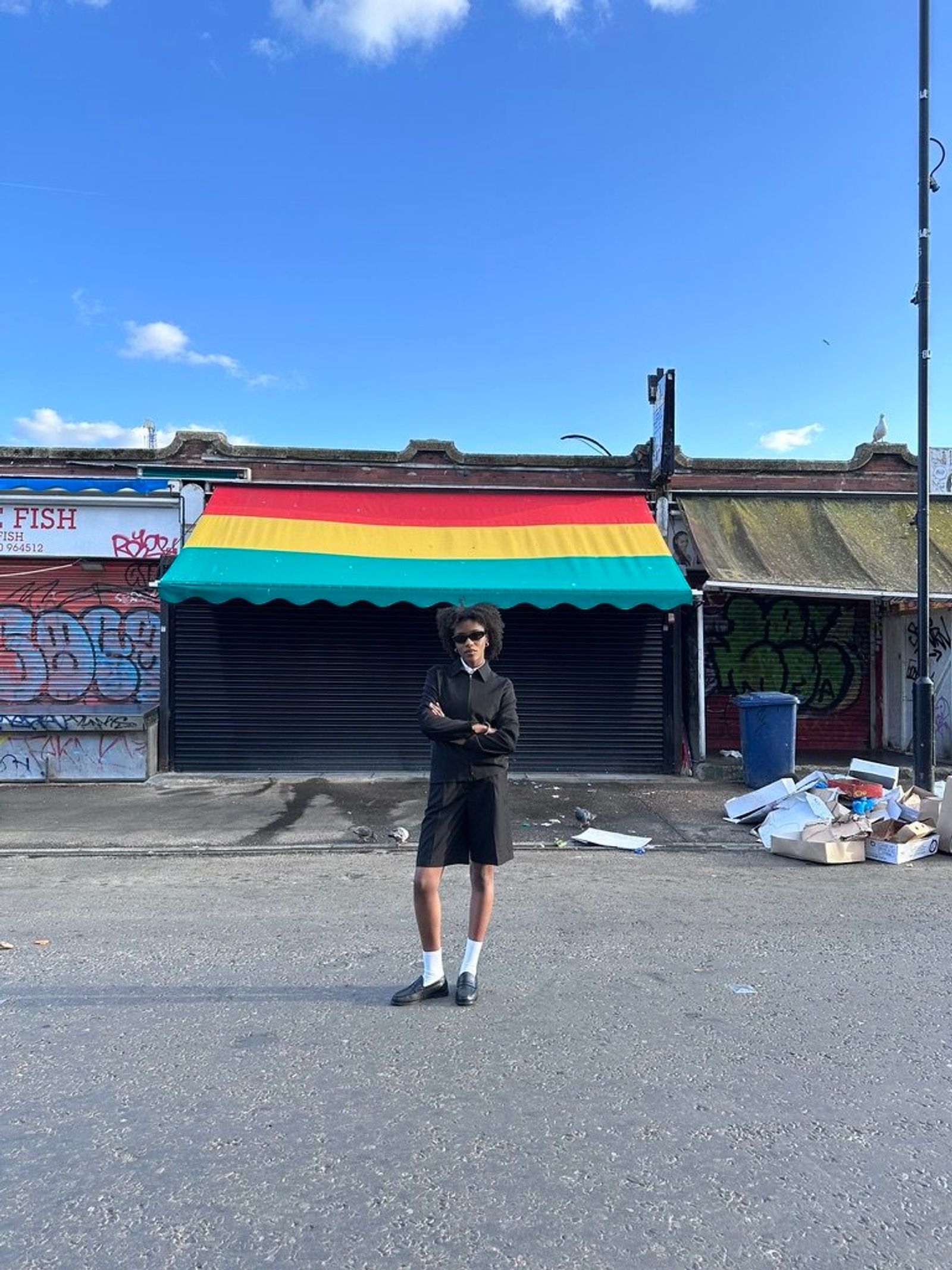 A person in a dark short suit, white socks and loafers stands with arms folded in front of a shuttered shop with a red, yellow and green awning.