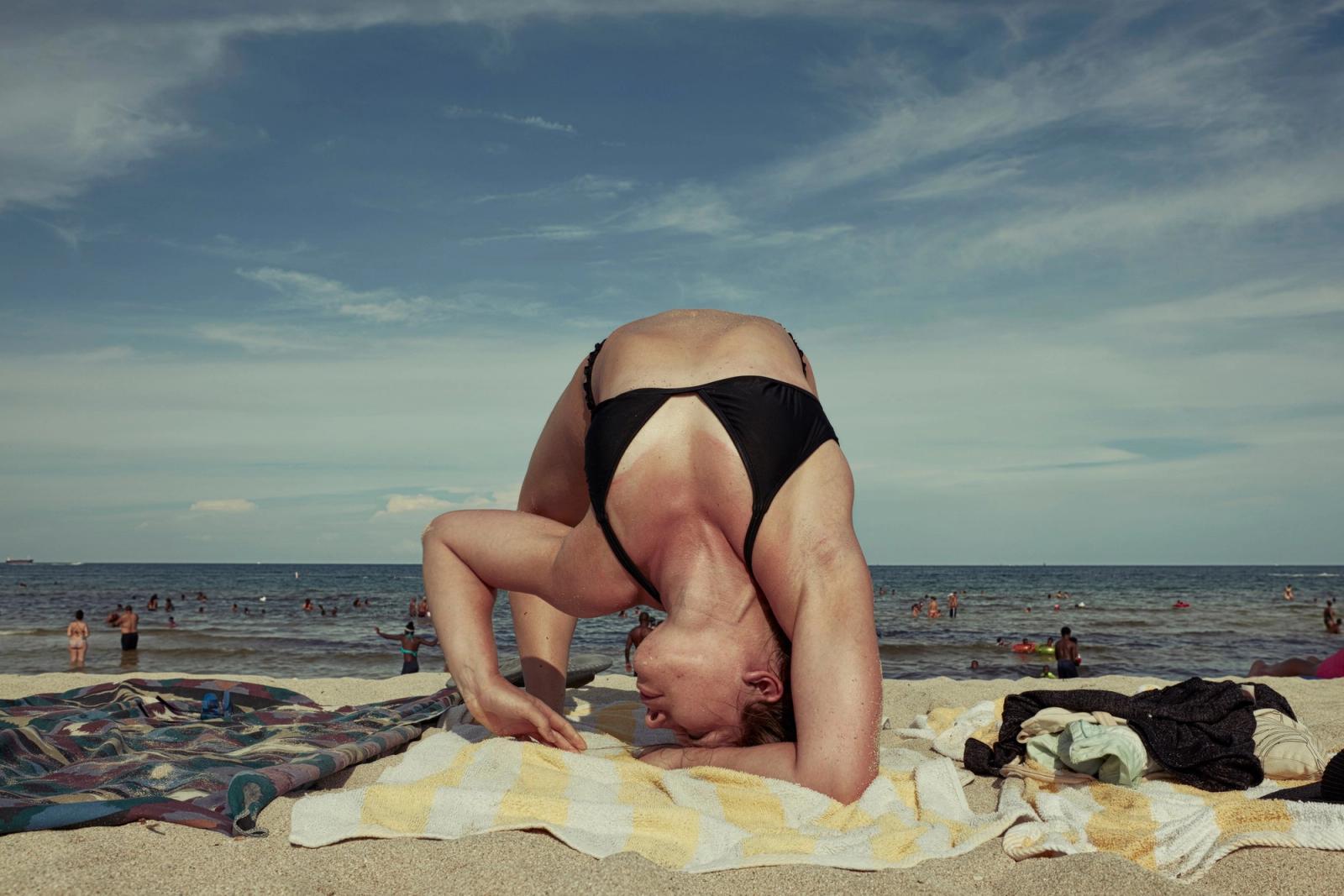 A woman in a black swimsuit balances in a headstand on a beach towel, with swimmers in the sea behind.