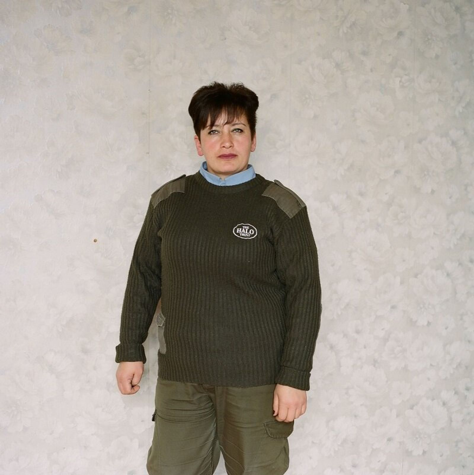 A woman with short dark hair in an olive sweater with a HALO Trust badge stands against a pale, textured wall.