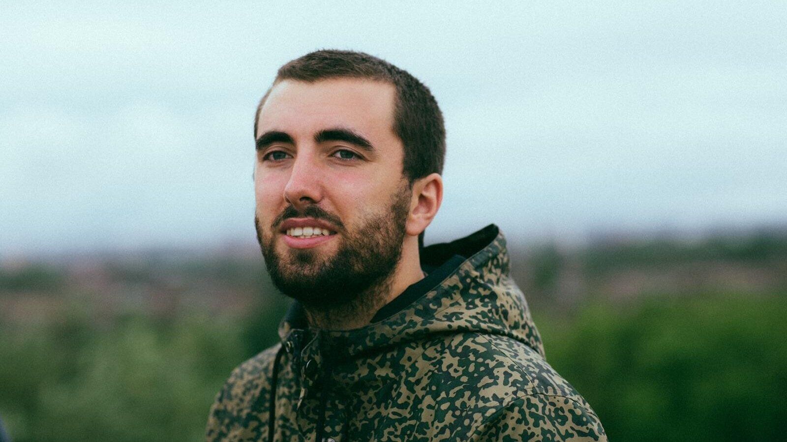 Milo Blake, smiling, with a short beard and a camouflage-print hoodie, against a cloudy sky and blurred green hills.