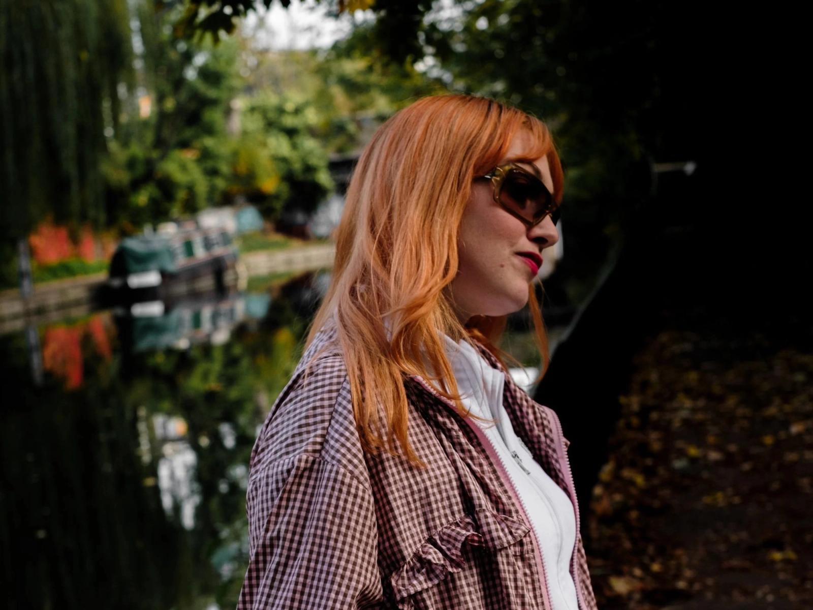Nancy June, with long orange hair, sunglasses and a checked jacket, beside a canal lined with narrowboats.