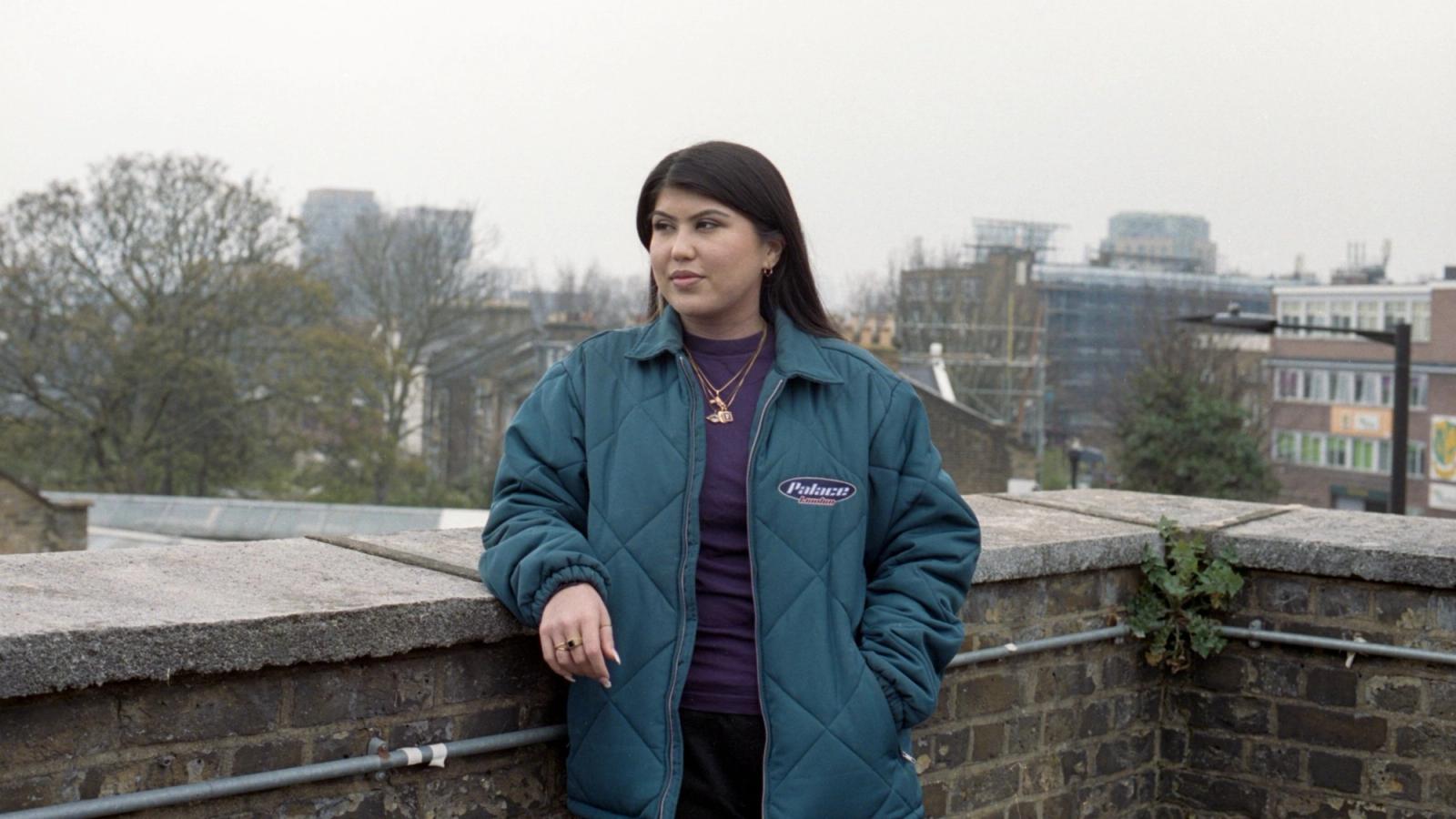 Freshta in a teal quilted jacket and gold necklace, leaning on a brick wall on a rooftop above city buildings.