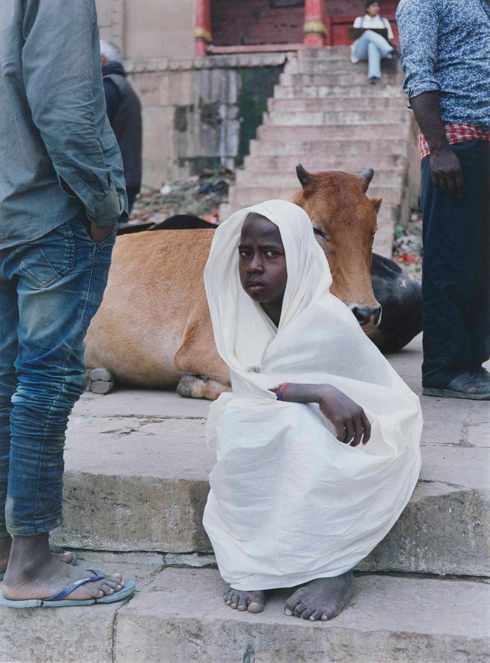 A child wrapped in white cloth sits on stone steps beside a resting cow, with people standing nearby.