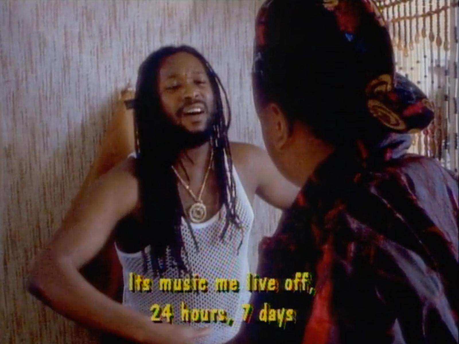 Film still from We The Ragamuffin: a man with long locs in a white mesh vest talks to a woman in a head wrap, with subtitles.
