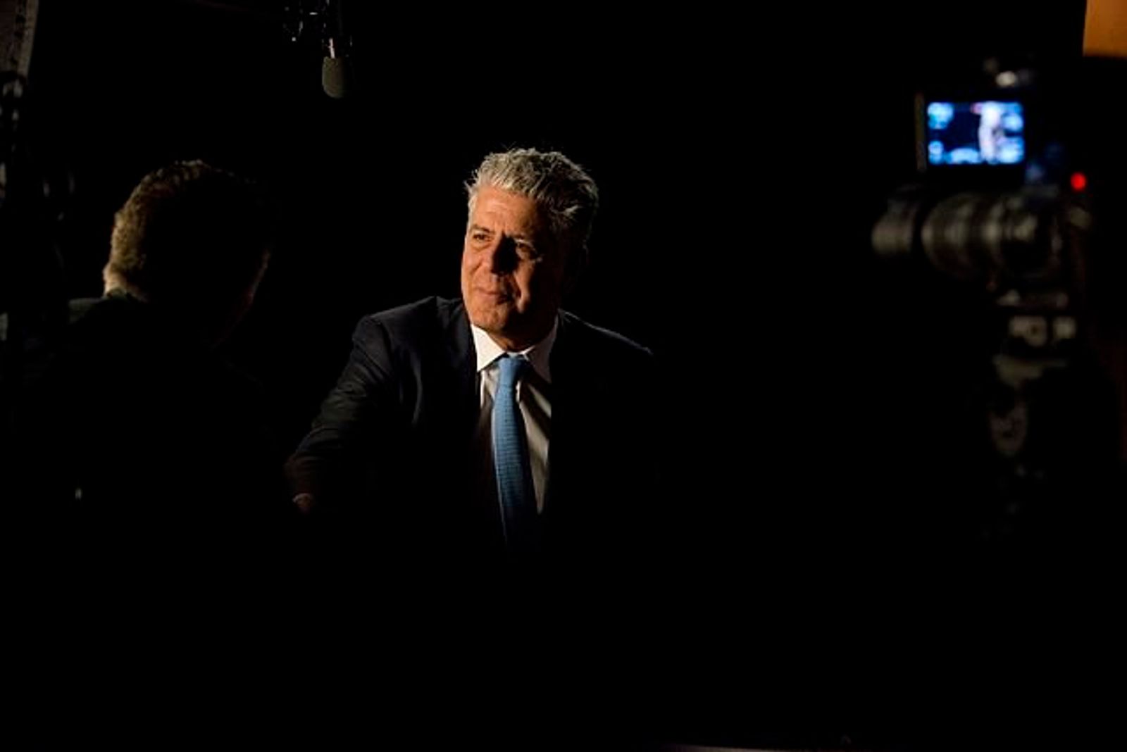 A grey-haired man in a dark suit and blue tie sits in a dark room, lit from the side, with a film camera beside him.