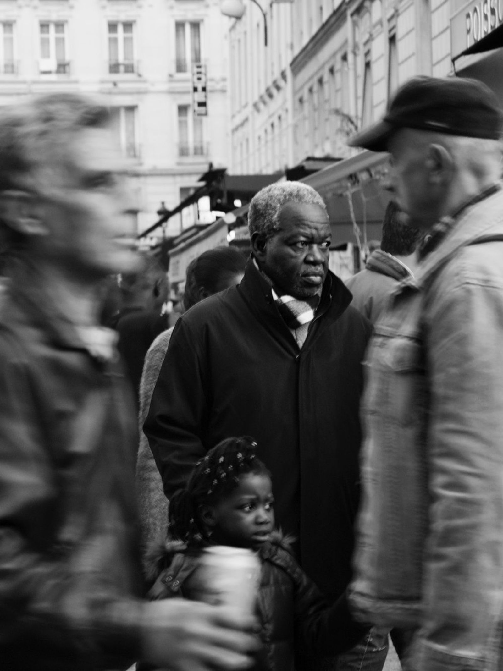 Black-and-white street photograph of an older man in a dark coat standing in a blurred crowd, with a small child beside him.