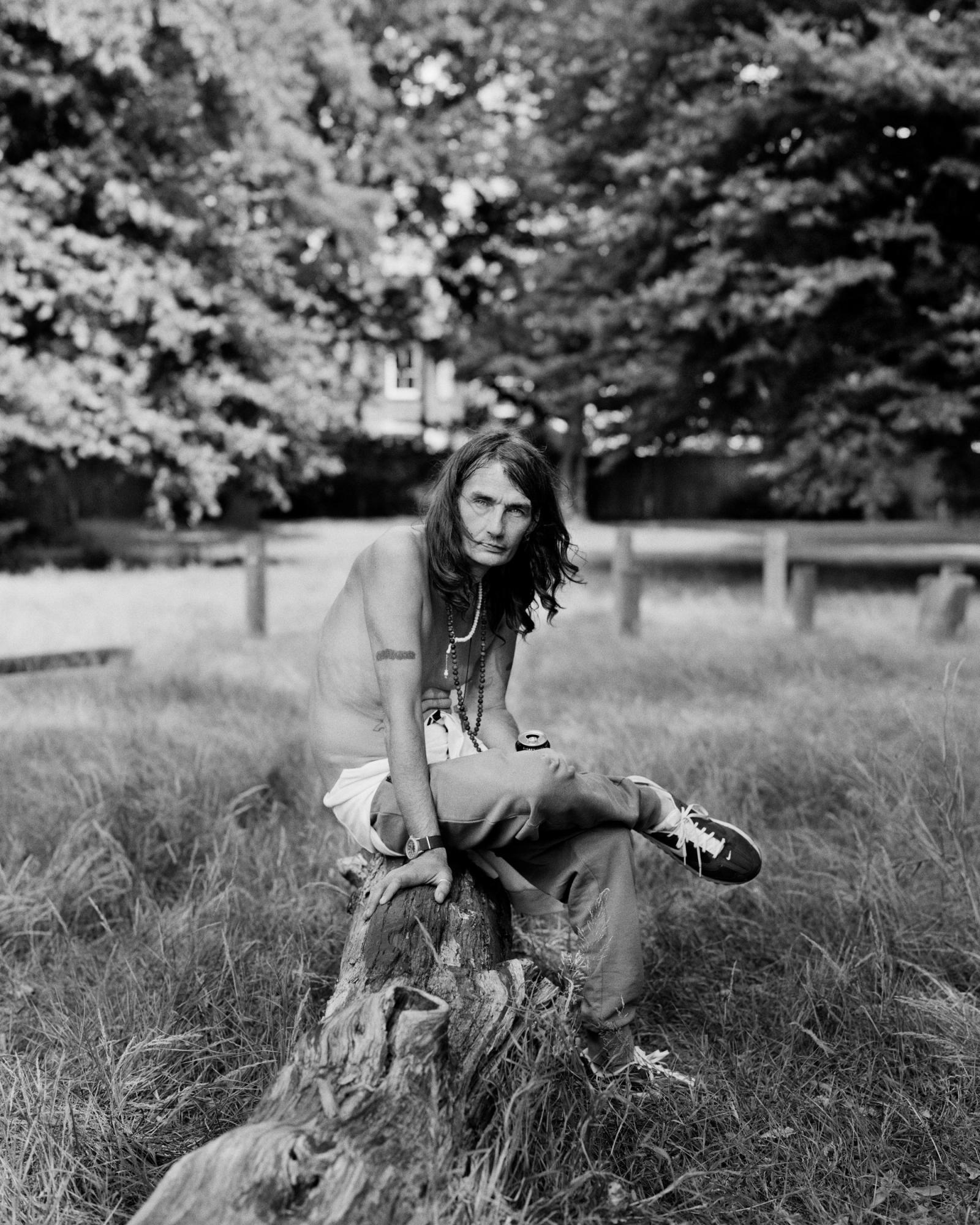 Black-and-white portrait of a long-haired man wearing bead necklaces, sitting shirtless on a fallen log in a park.