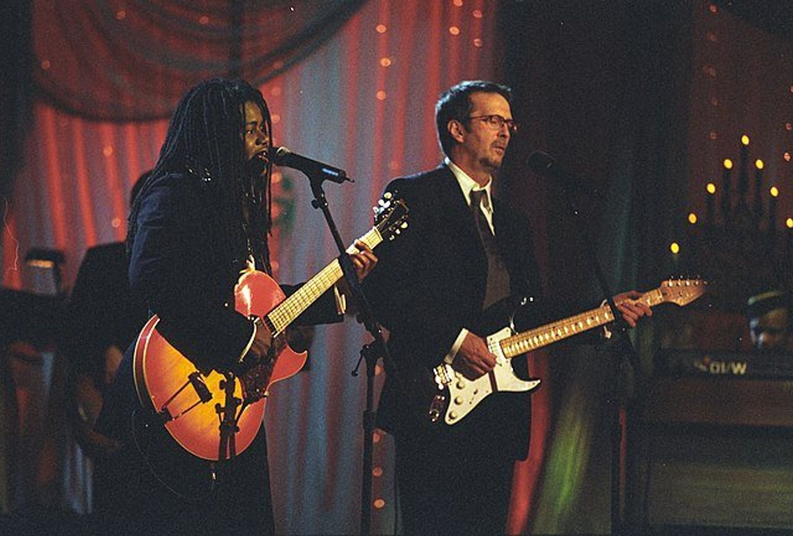 Two men on stage playing guitars: one with long locs sings at a microphone, the other in a dark suit plays an electric guitar.