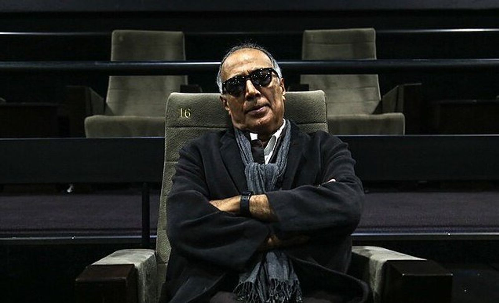 A man in dark sunglasses and a dark jacket sits with his arms crossed in a cinema seat, with empty rows behind him.