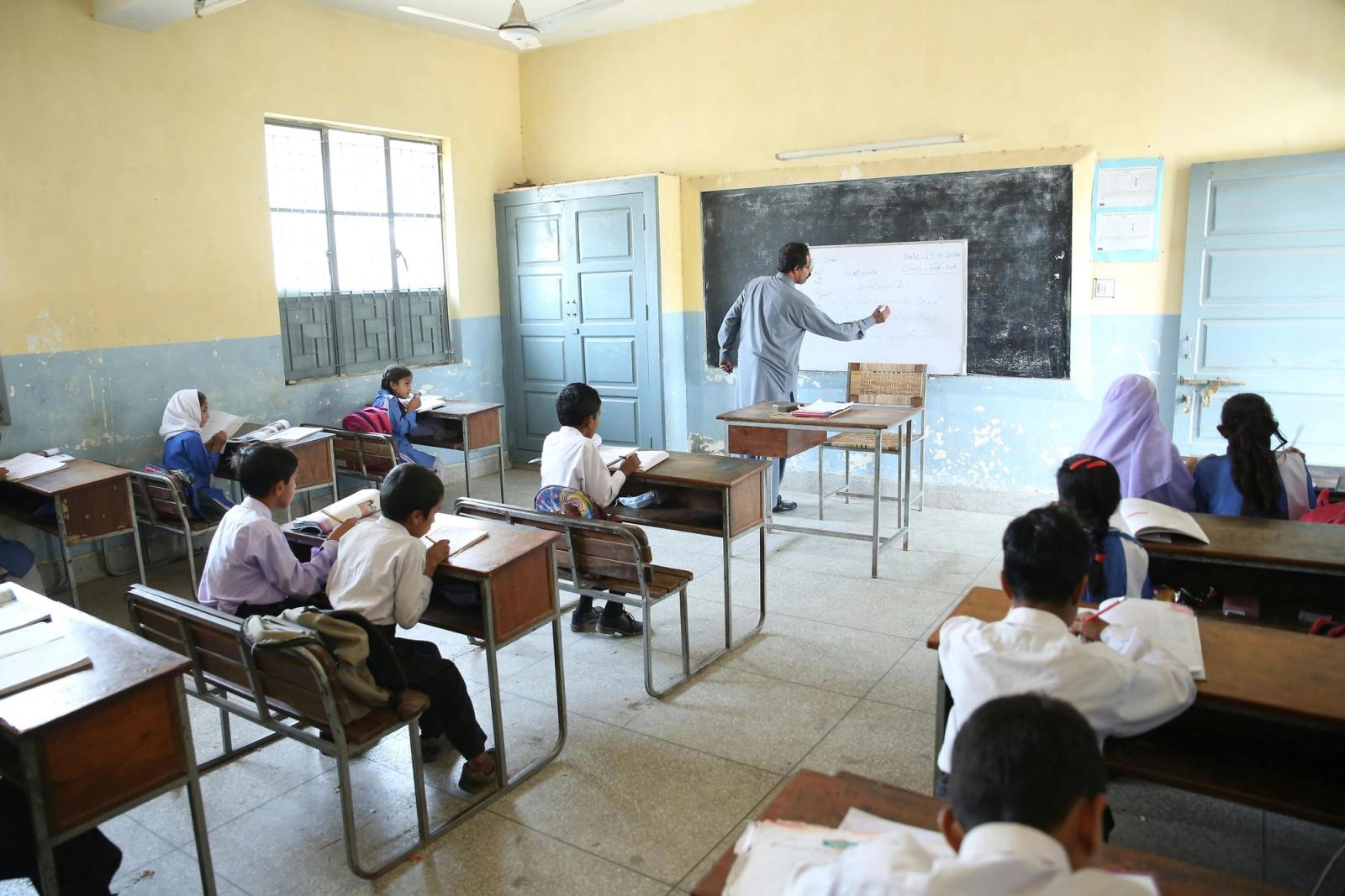 A teacher writes on a whiteboard in a yellow-walled classroom as children sit at wooden desks.