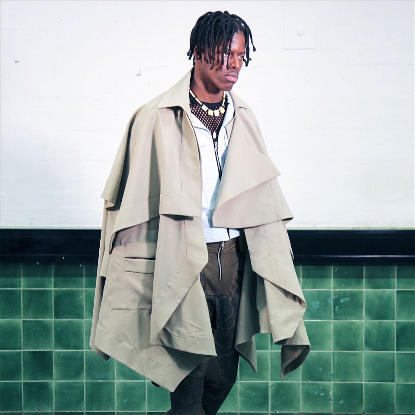 A model with locs wears a layered beige cape over a pale shirt and a mesh necklace, in front of a green-tiled wall.