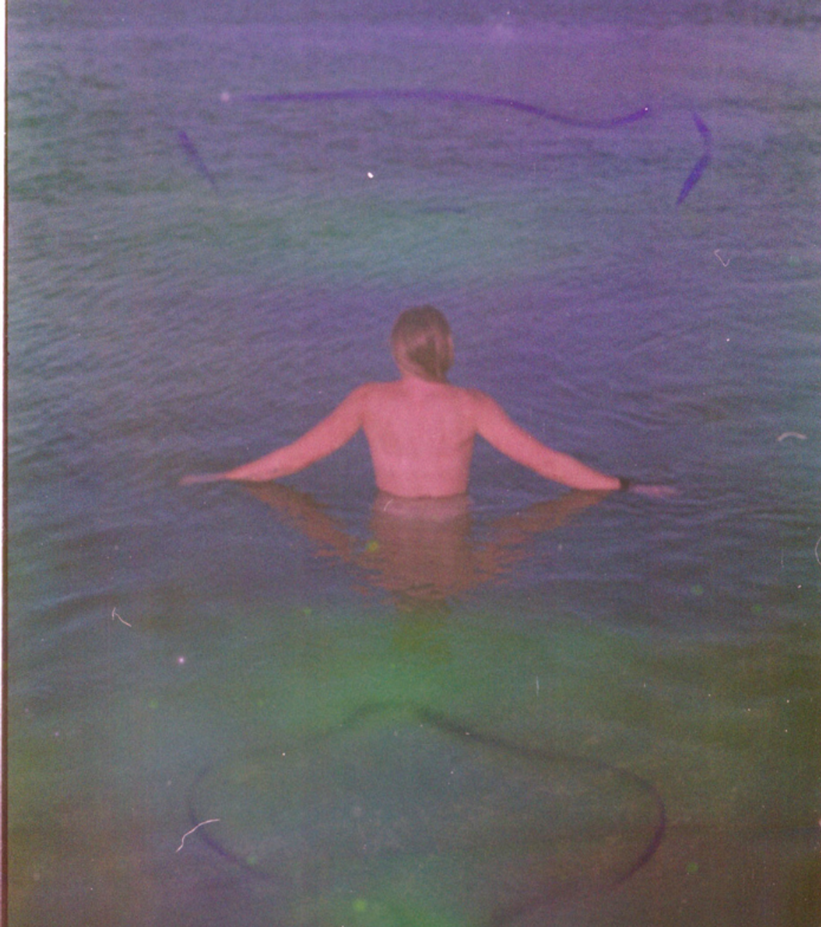 Damaged film photograph of a person standing waist-deep in water, seen from behind with arms outstretched, tinted purple and green.