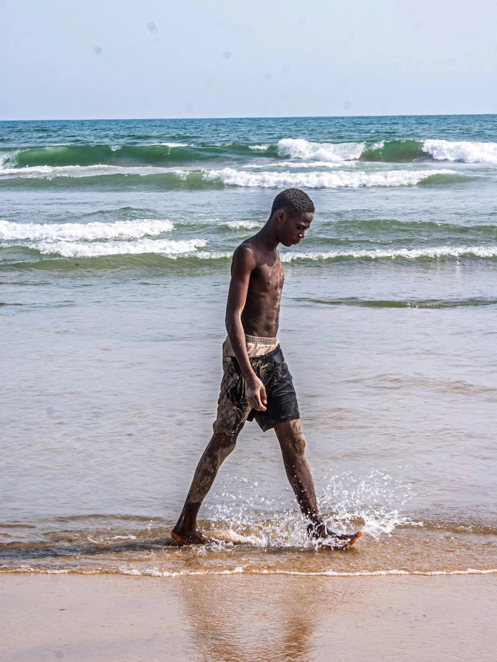 A boy in dark shorts walks barefoot through shallow surf, waves breaking behind him.