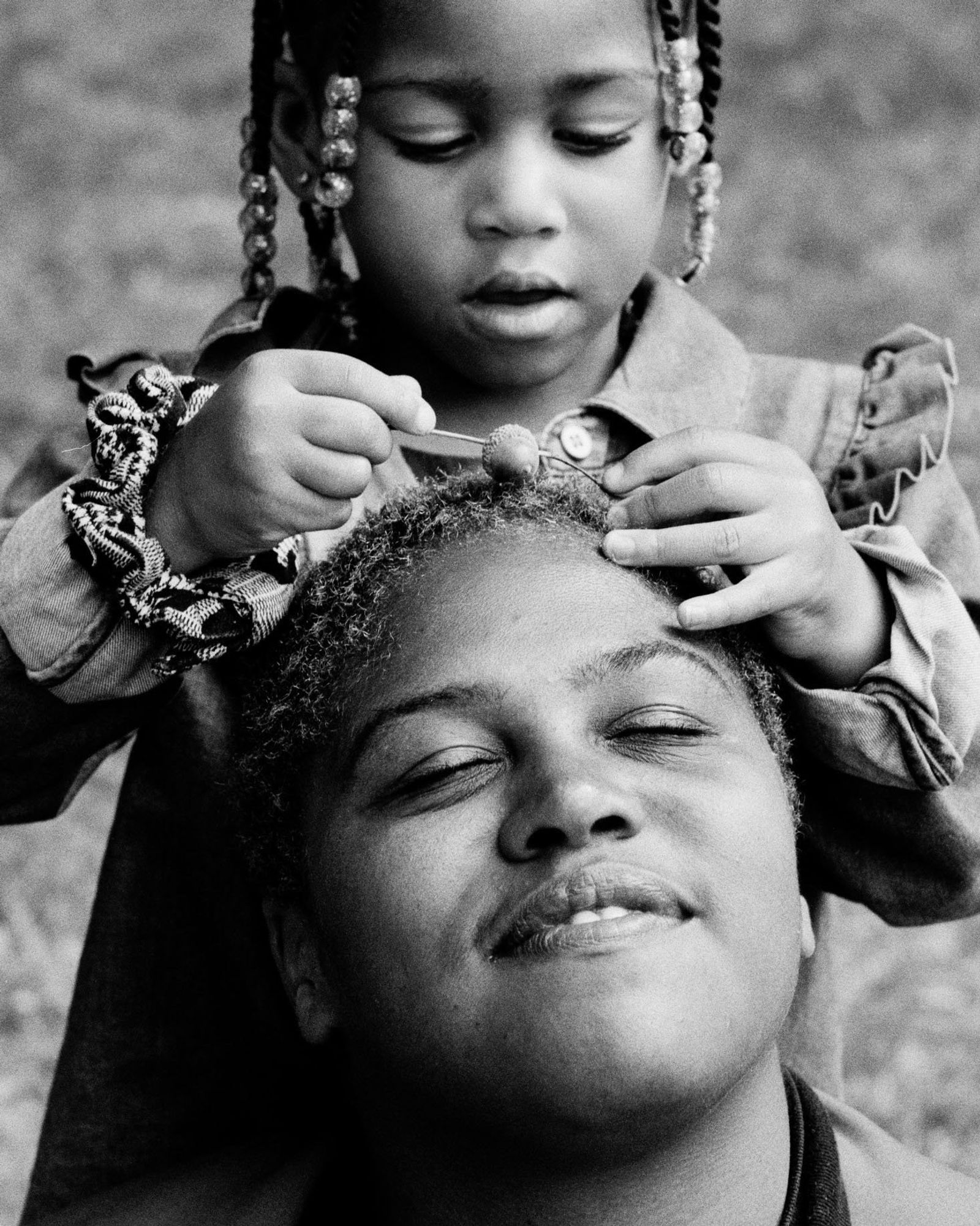 Black-and-white photo of a young girl with beaded braids placing a bead in a woman's short hair; the woman smiles with her eyes closed.