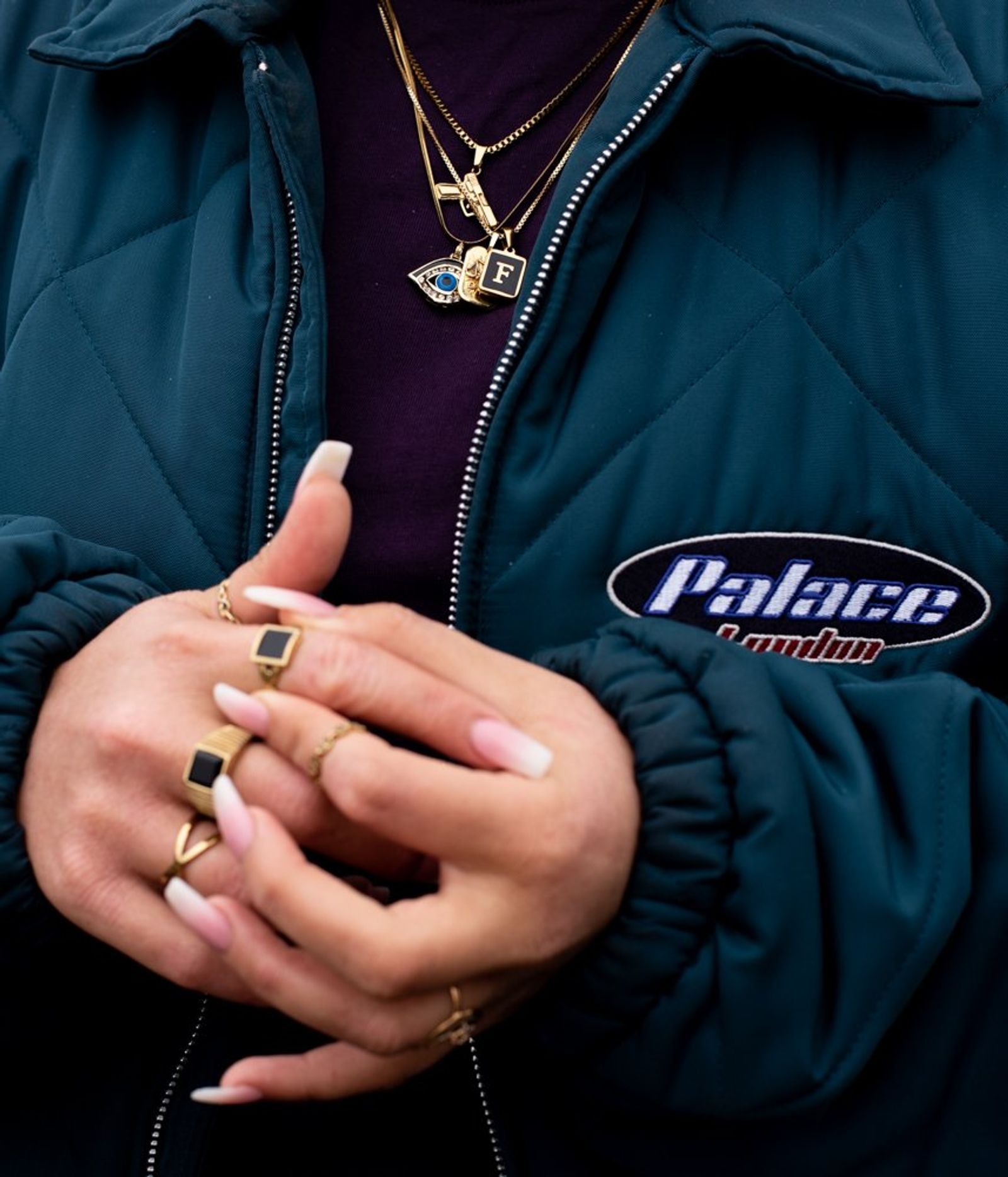 Close-up of Freshta's hands with gold rings and long nails, over a teal quilted jacket and layered gold necklaces.