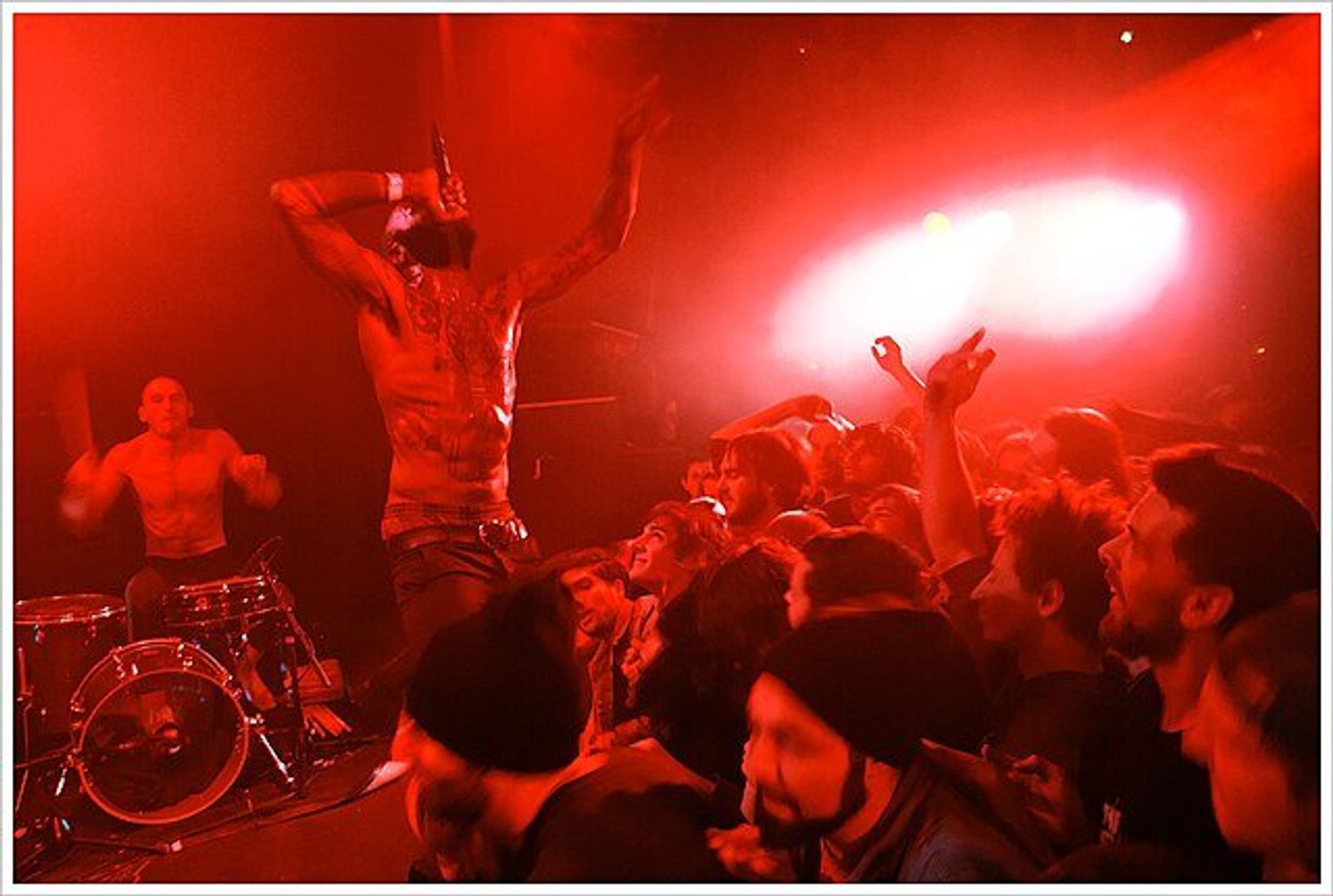 A shirtless performer with raised arms stands on a red-lit stage above an excited crowd, with a drummer behind.