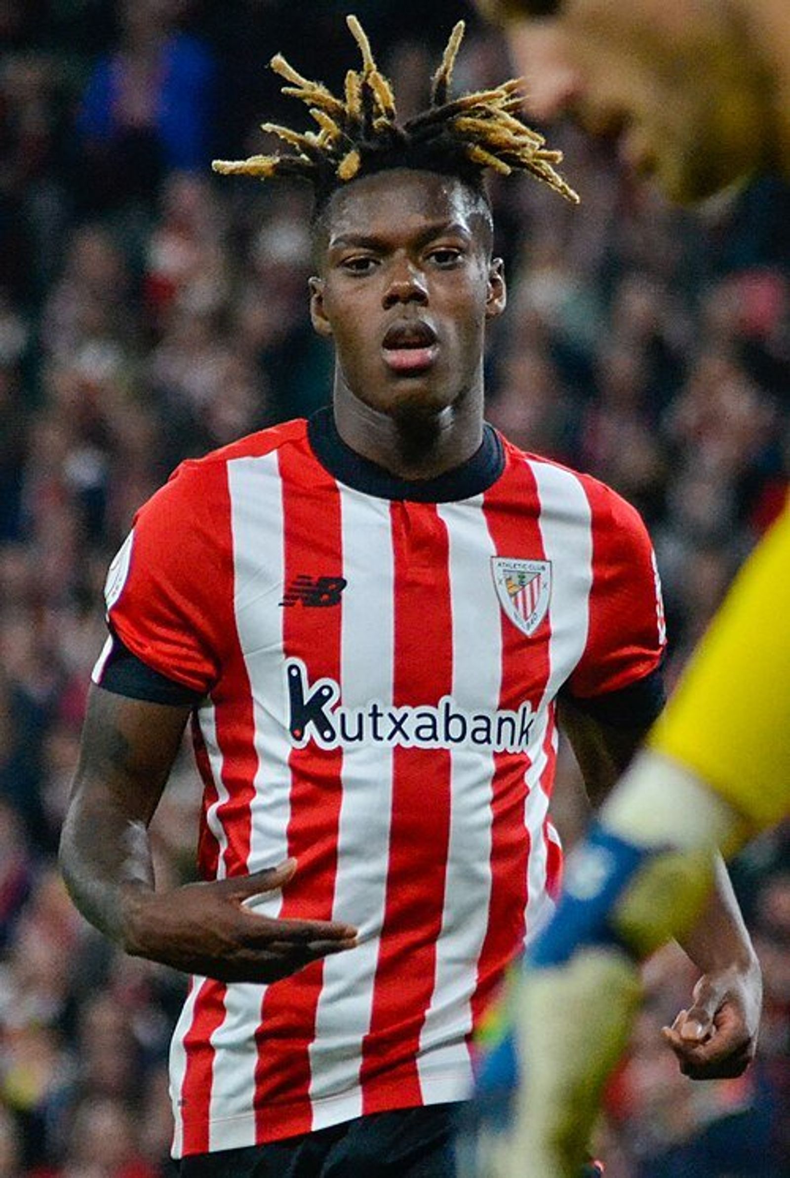 A footballer in a red and white striped Athletic Club shirt, with blond-tipped locs, looks ahead during a match, with a crowd behind.