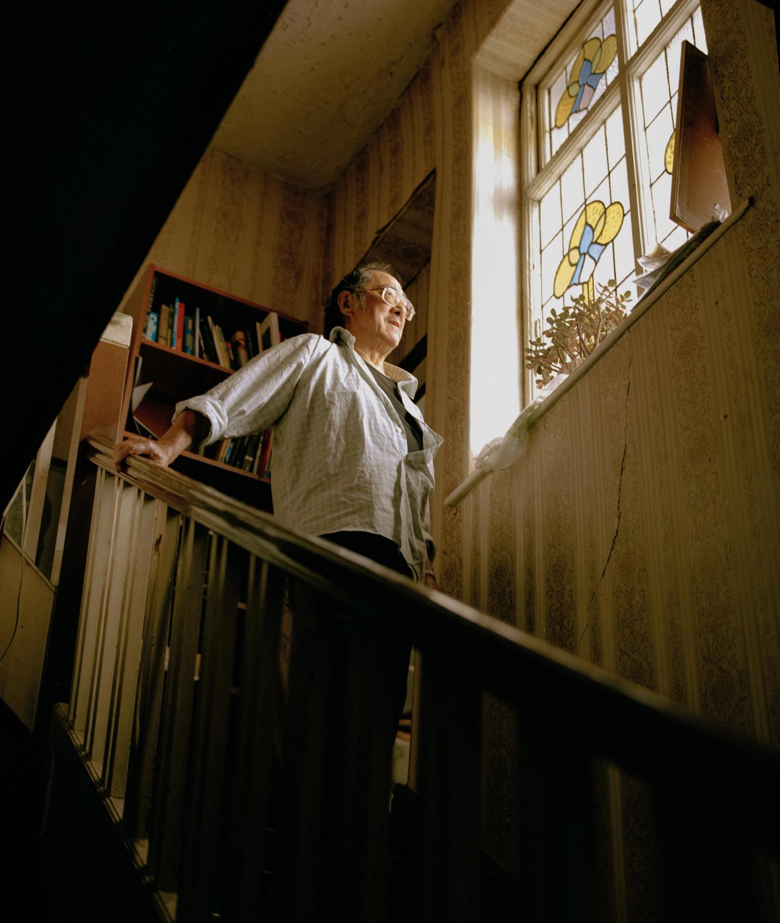 An older man stands at the top of a staircase beside a stained-glass window, looking off to one side.
