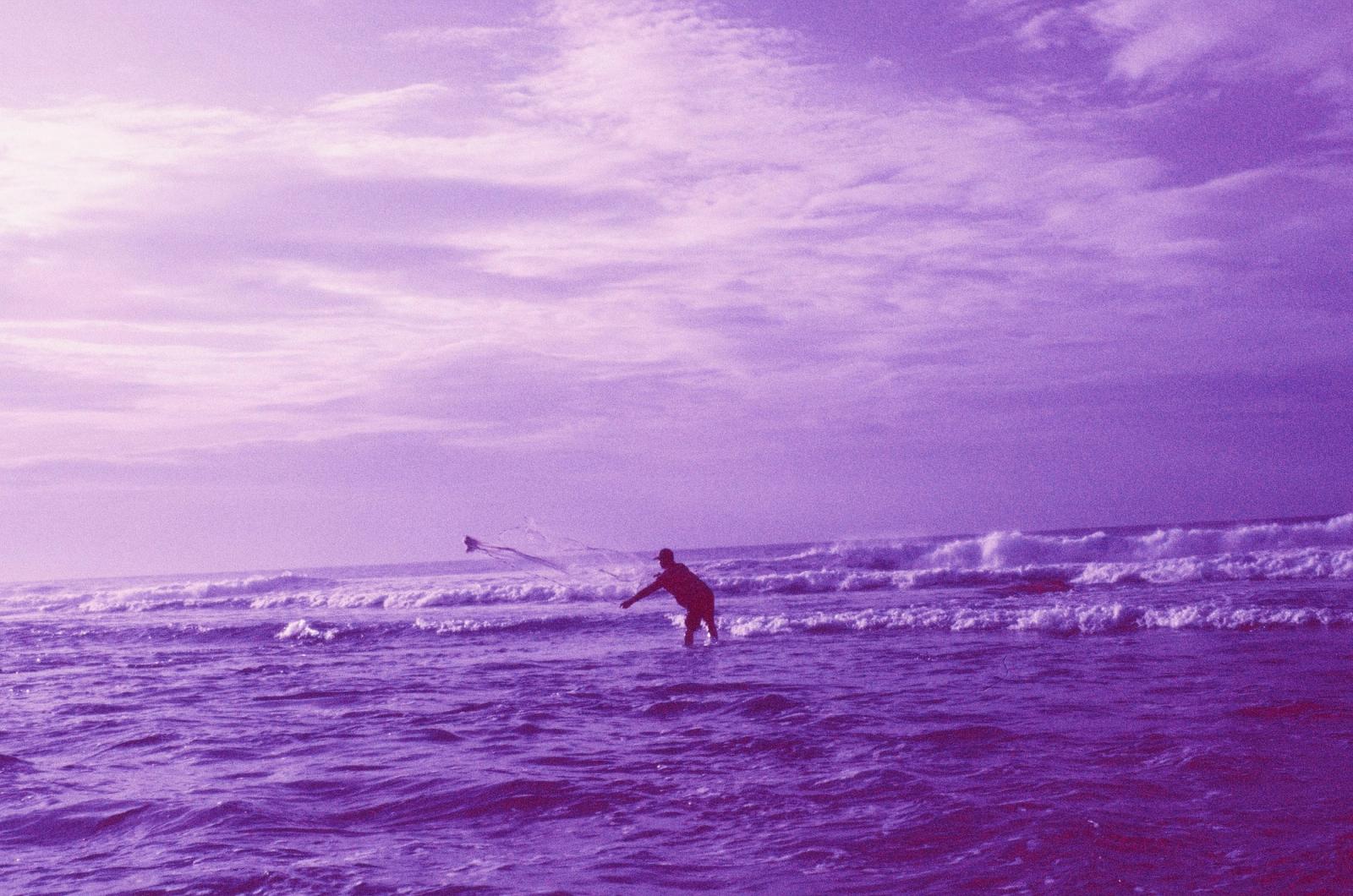 A fisher stands in rough surf casting a net, photographed on purple-toned Aerochrome film.